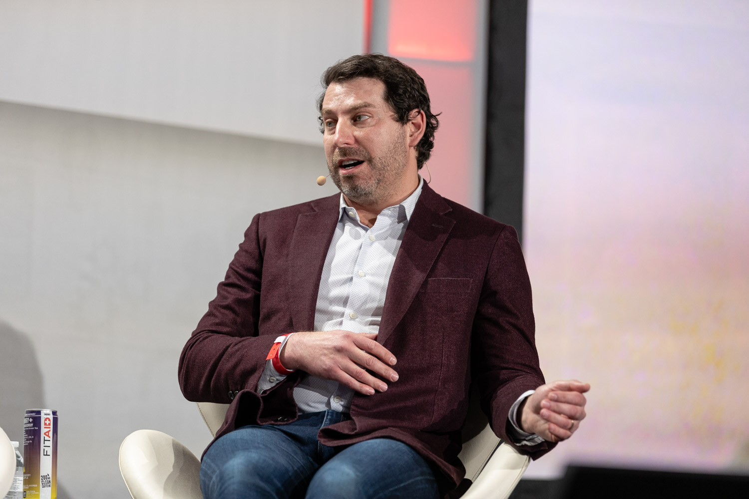 A dark-haired man in a burgundy blazer and jeans sits and gestures with one open hand, with cans on a small table beside him.