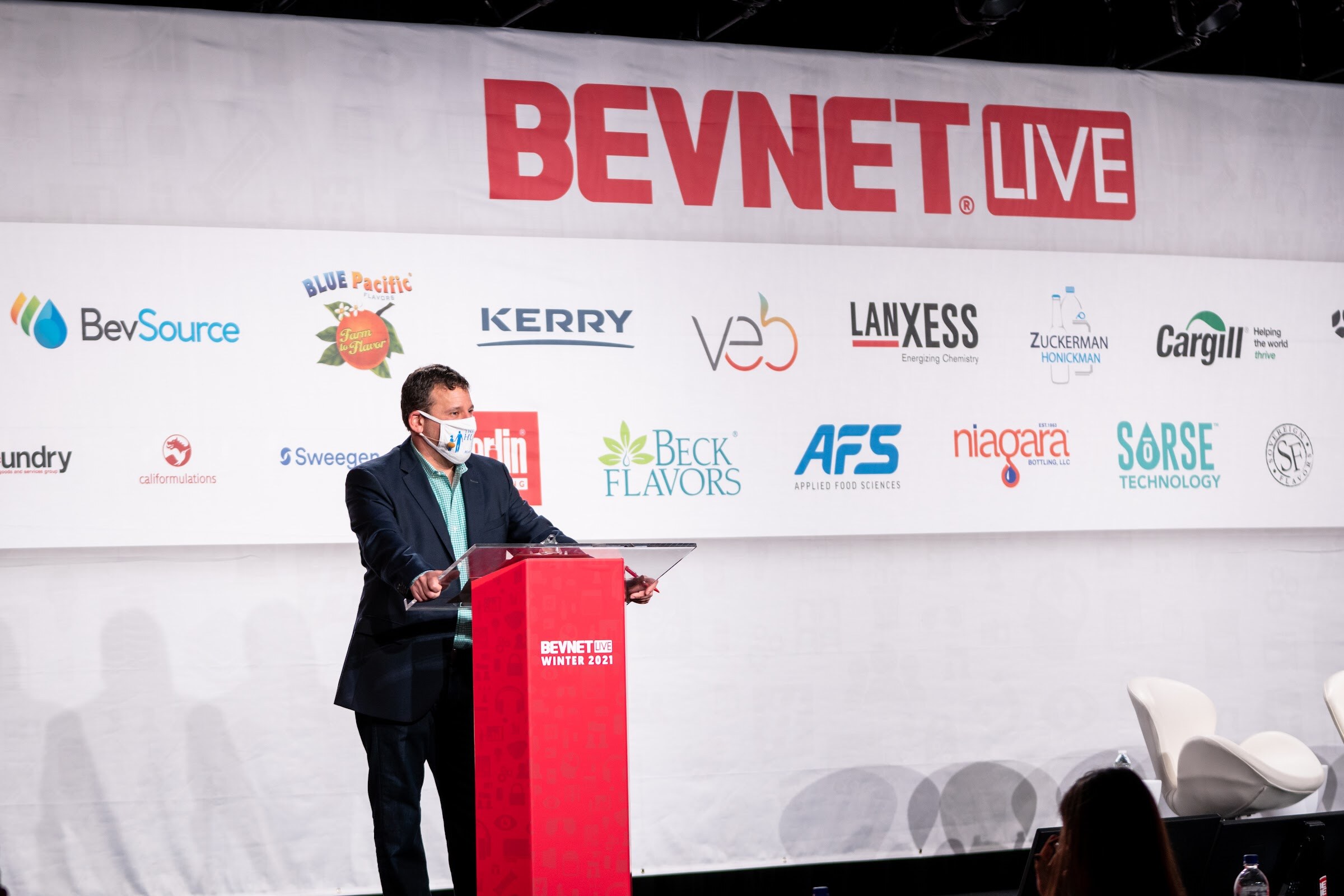 A man in a navy blazer and face mask stands at a red podium on a stage in front of a BevNET Live banner and sponsor logos.