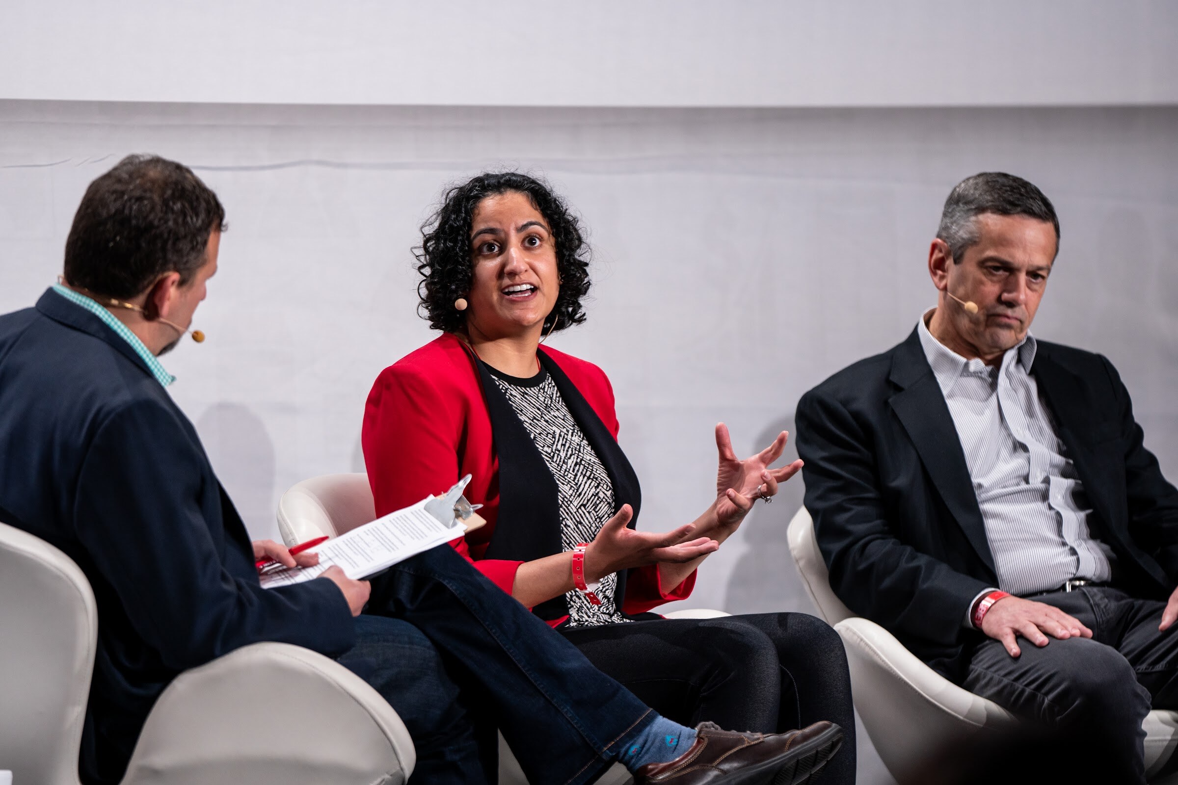 A woman with curly dark hair in a red blazer gestures with both hands while a moderator and another panelist listen.