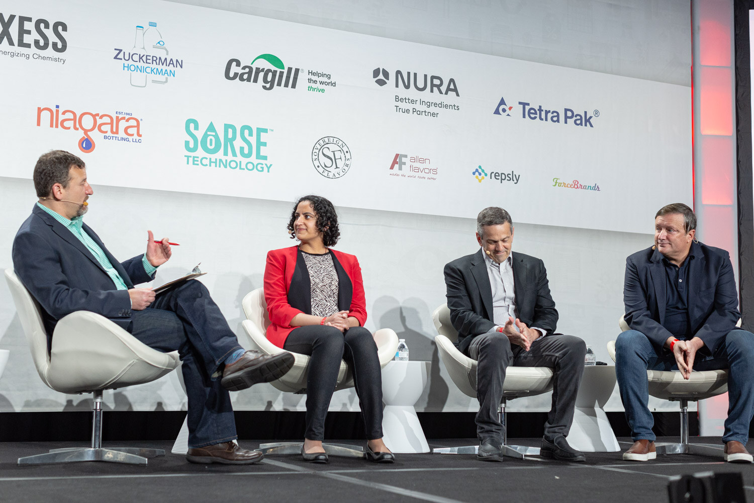 Four people seated in white chairs on stage, with the moderator at left holding papers and a panelist in a red blazer next to him.
