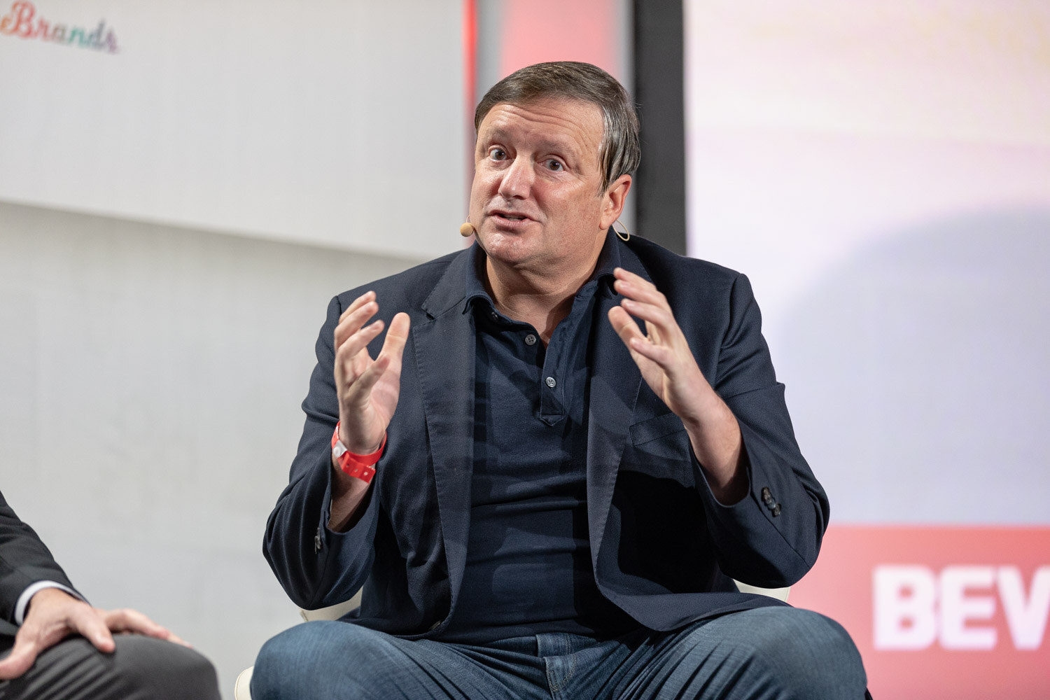 A man in a dark blazer and navy shirt gestures with both hands raised in front of a white backdrop with a red BEV graphic.