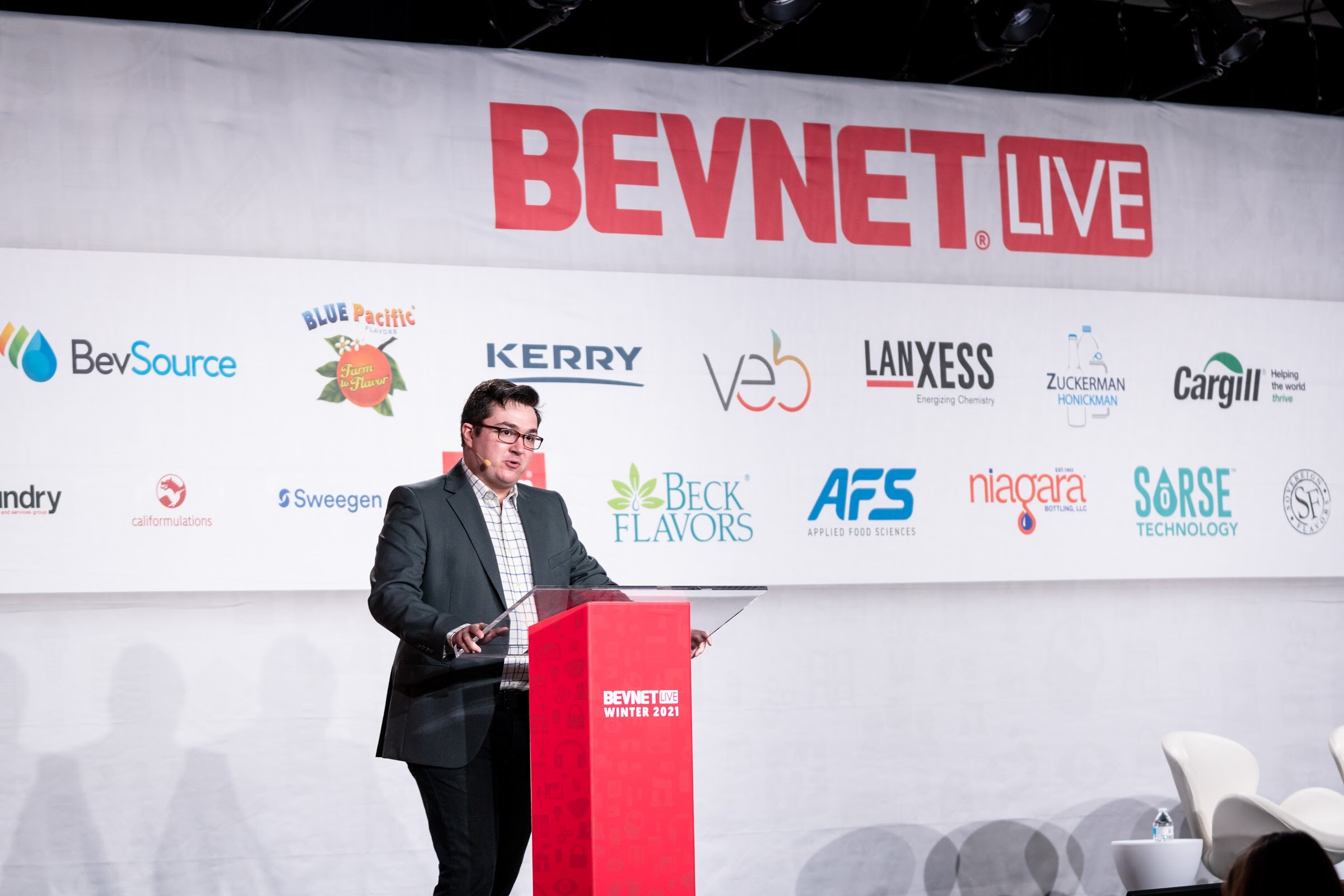 A man in glasses and a dark blazer stands at a red podium on a stage below a BevNET Live banner and sponsor logos.
