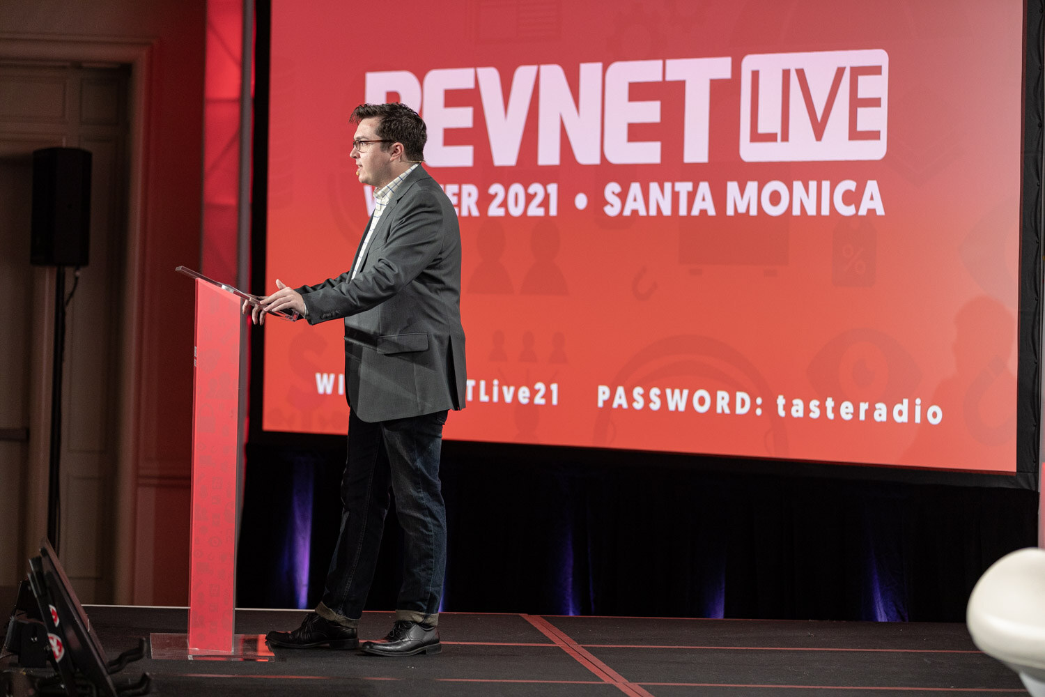 A man in a gray blazer stands at a red podium looking at a laptop in front of a large red screen reading BevNET Live Winter 2021 Santa Monica.