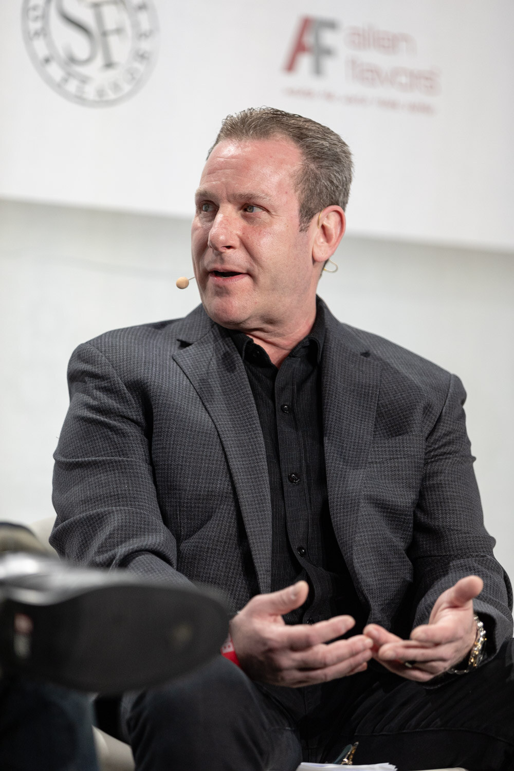 A man in a gray blazer and black shirt sits in a white chair gesturing with both hands in front of a white sponsor wall.