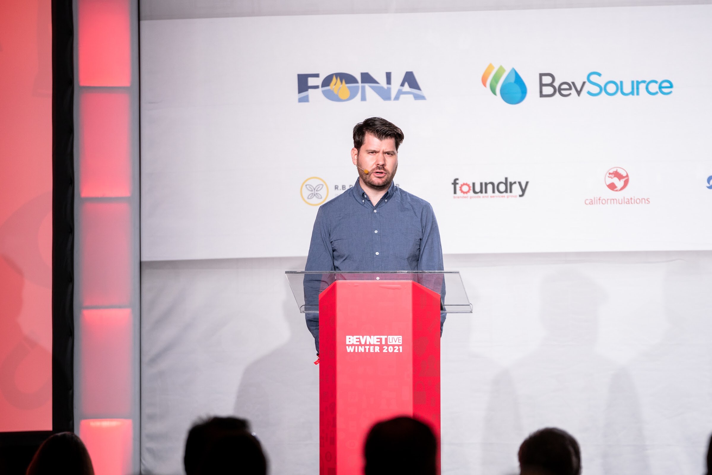 A man in a blue button-up shirt stands at a clear lectern in front of a white wall with FONA, BevSource and Foundry logos.