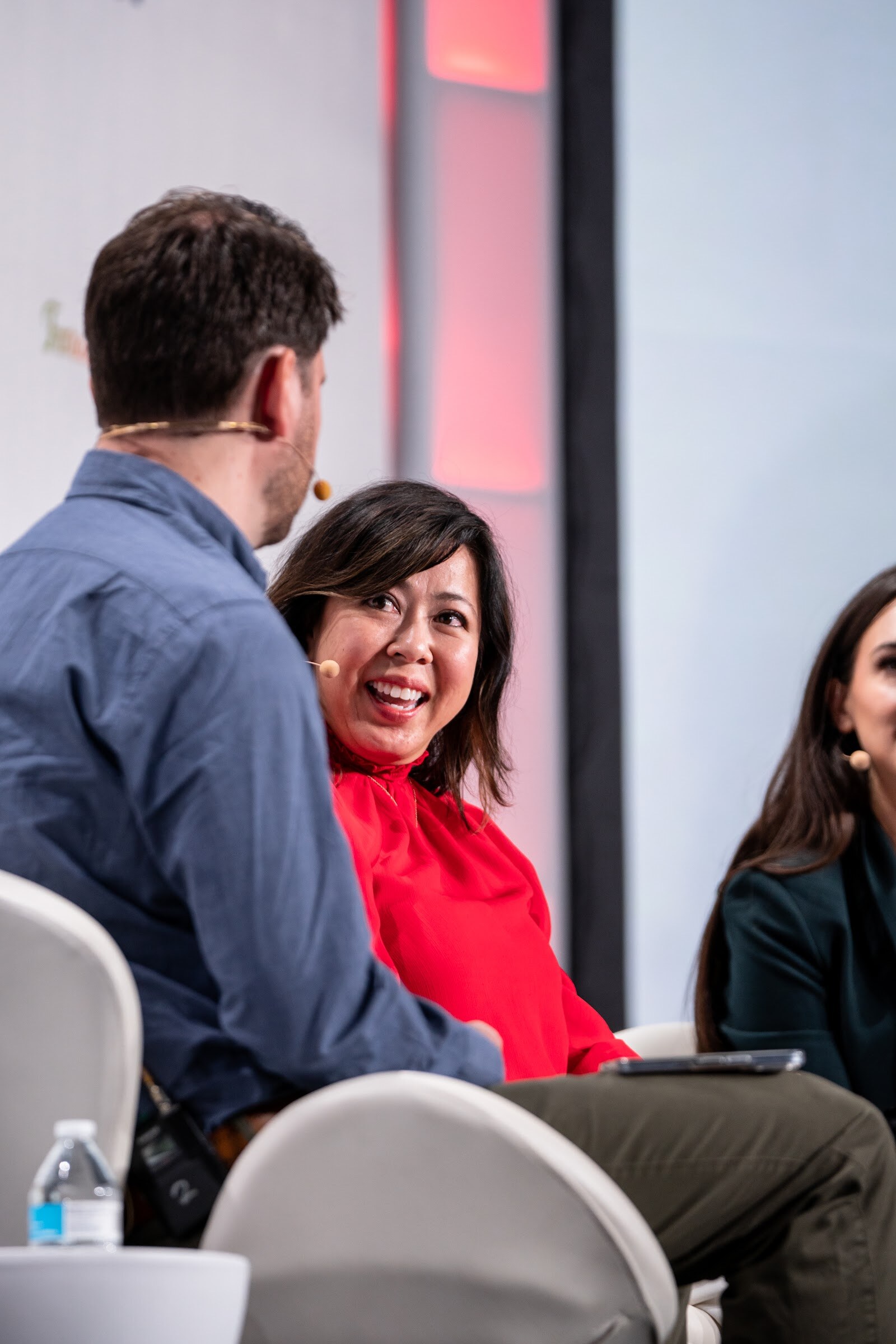 A smiling woman in a red top with a headset microphone sits between a man in a blue shirt seen from behind and a woman in a green blazer at the right edge.