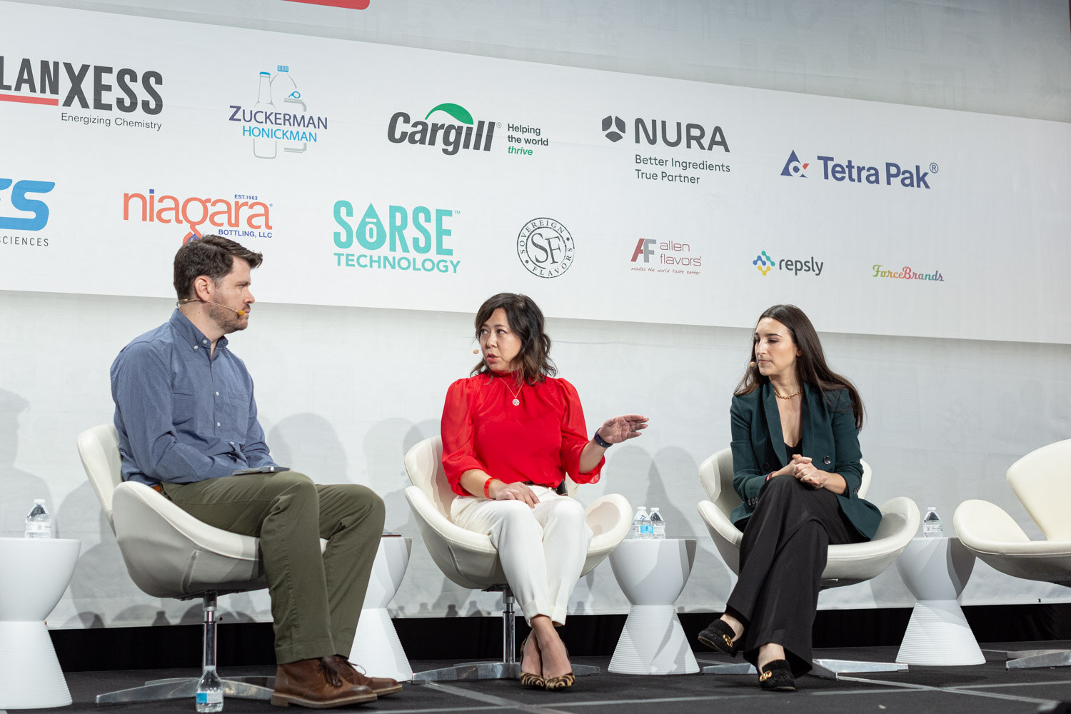A man in a blue shirt and two women, one in a red blouse and one in a dark green blazer, sit in white chairs in front of a sponsor wall.