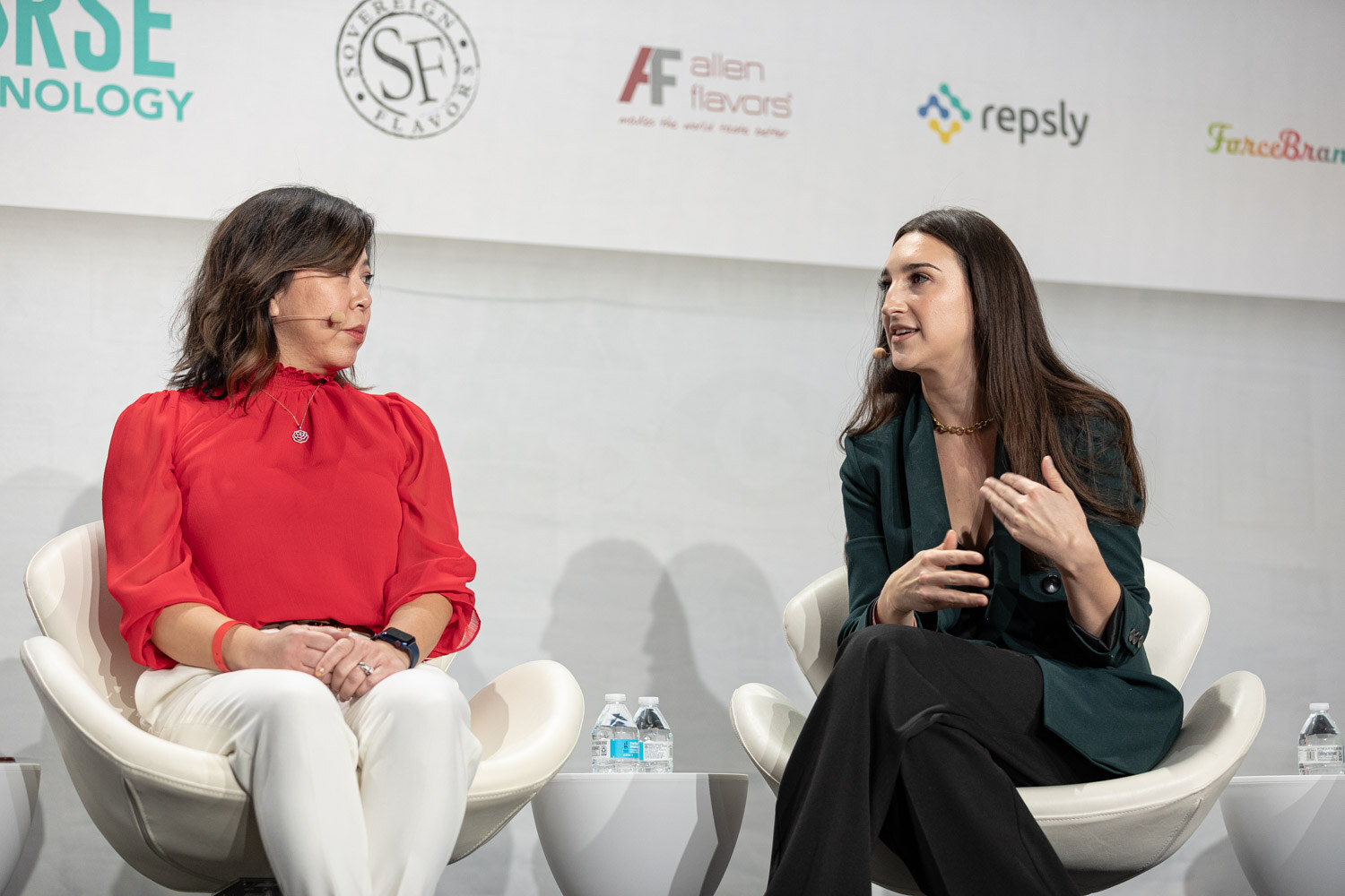 A woman in a red blouse and a woman in a dark green blazer sit in white chairs and talk, with water bottles on small tables.