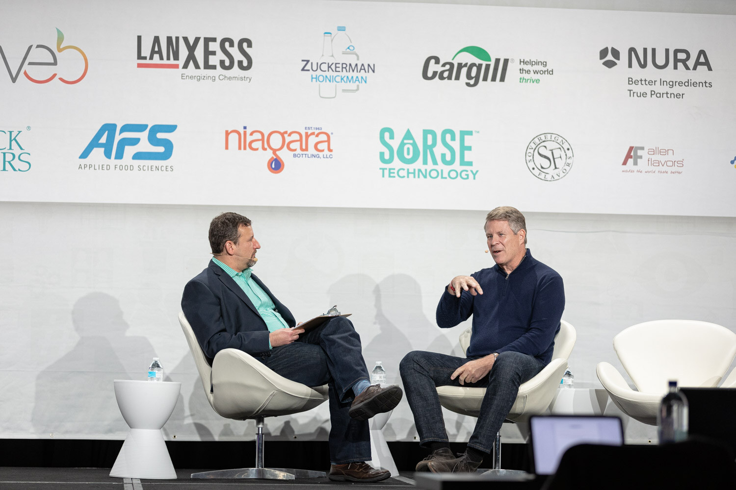 Two men seated in white chairs on stage in front of a sponsor wall, one in a navy jacket with a notepad and one in a navy quarter-zip sweater gesturing.