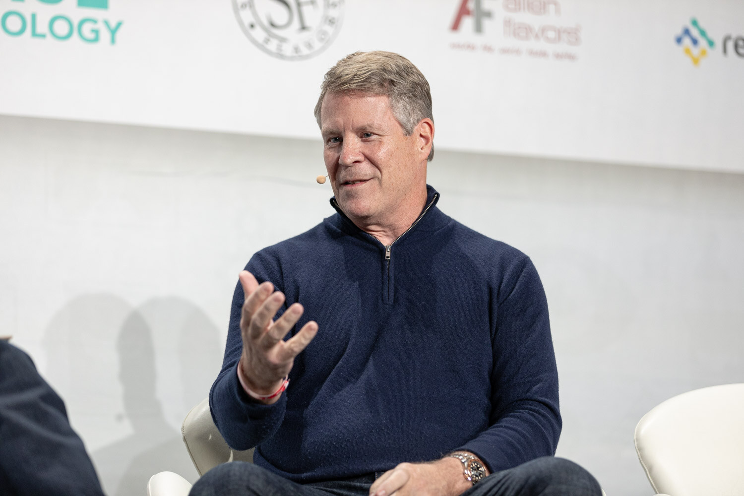 A man with swept gray-blond hair in a navy quarter-zip sweater raises one open hand while he talks.