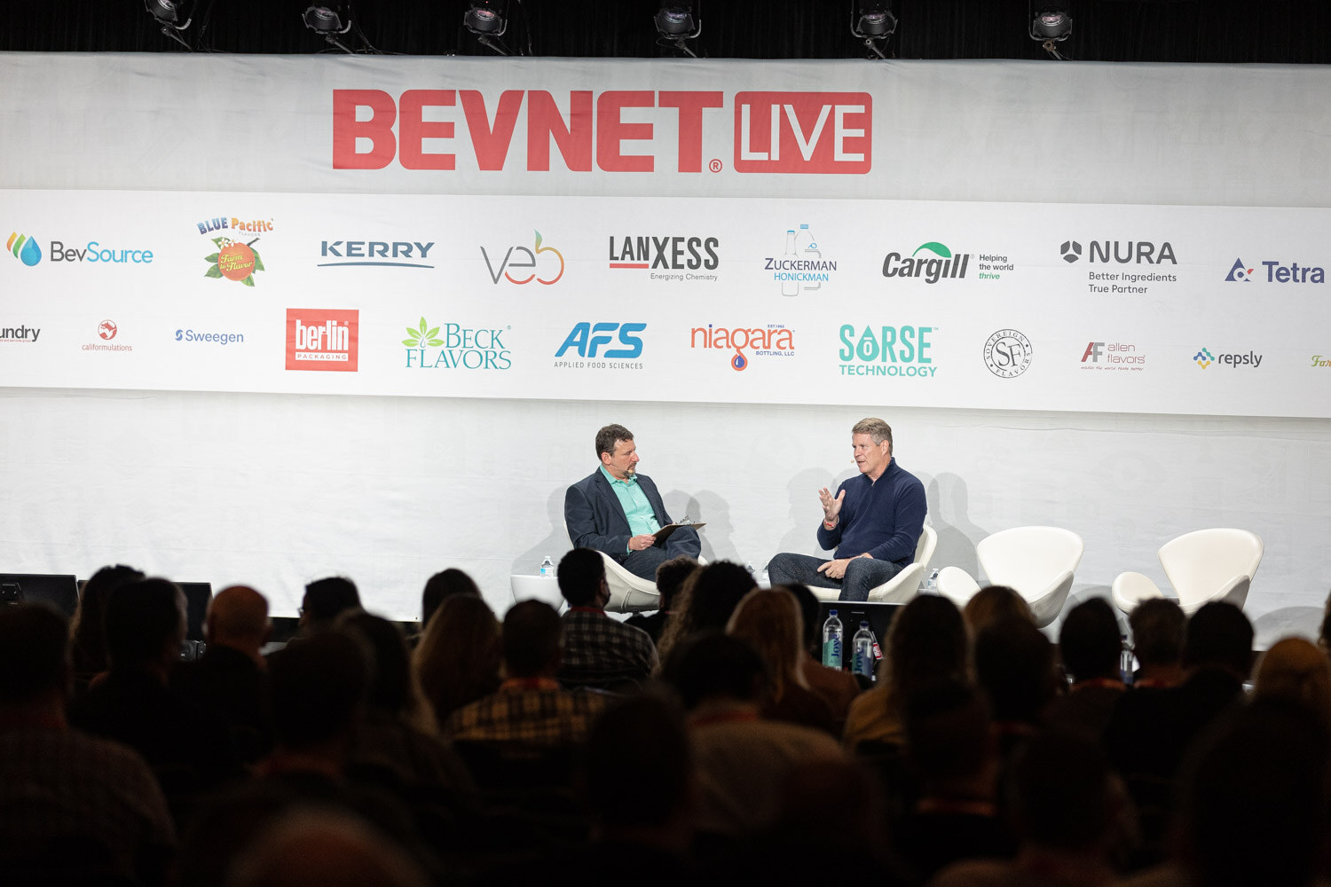 A wide view of two men in conversation on a stage below a BevNET Live banner, with the silhouetted audience in the foreground.