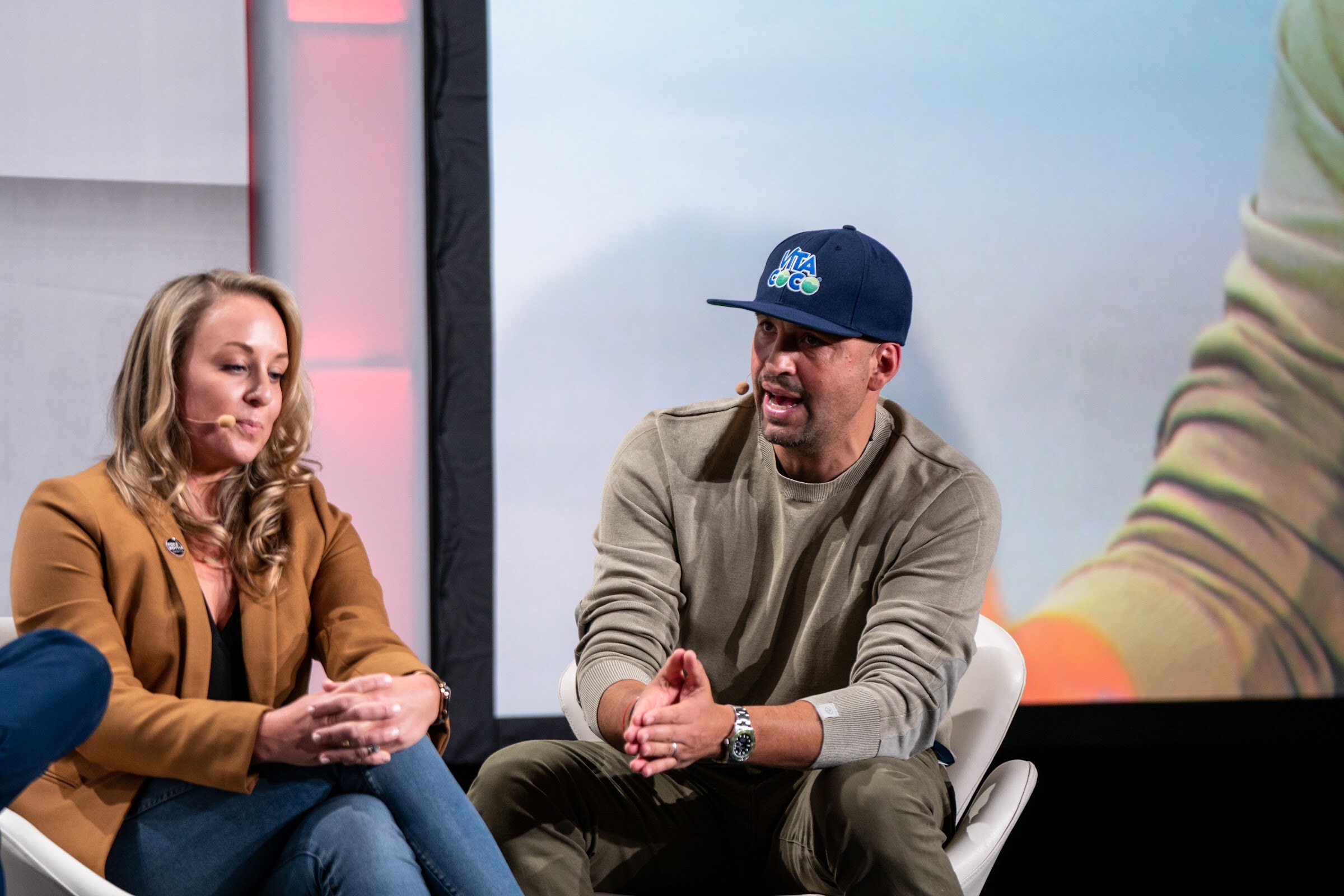A woman with long wavy hair in a camel blazer and a man in a navy cap and olive sweater sit side by side and talk.
