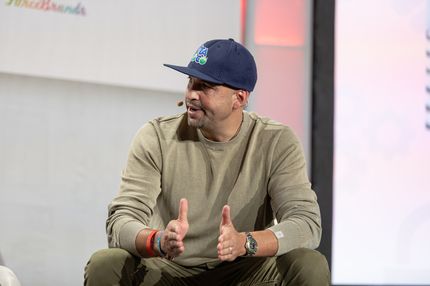 A man in a navy Vita Coco cap and olive sweatshirt sits leaning forward with his hands apart while speaking.