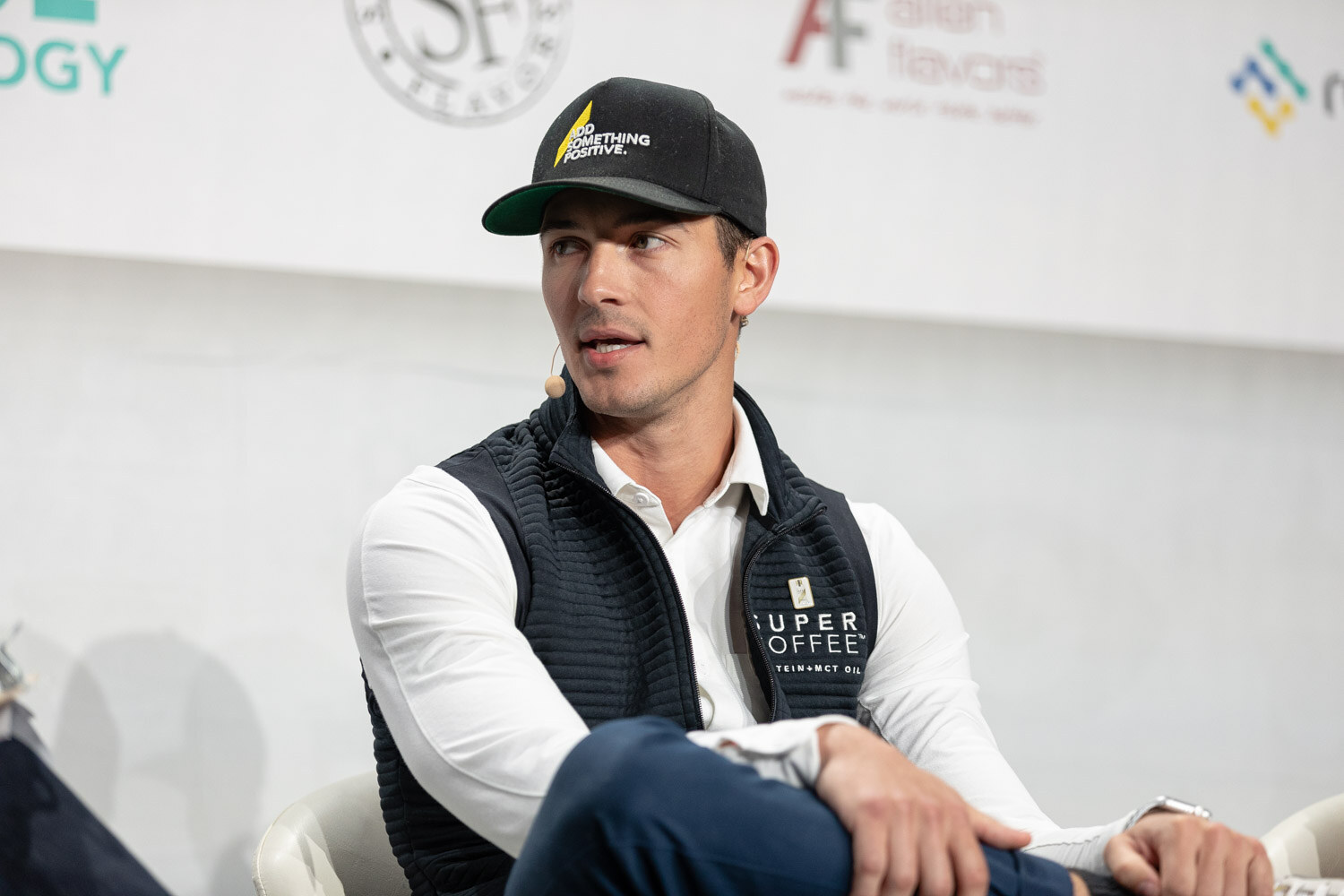 A man in a black cap and a navy Super Coffee vest over a white shirt sits with one leg crossed, looking to his left.