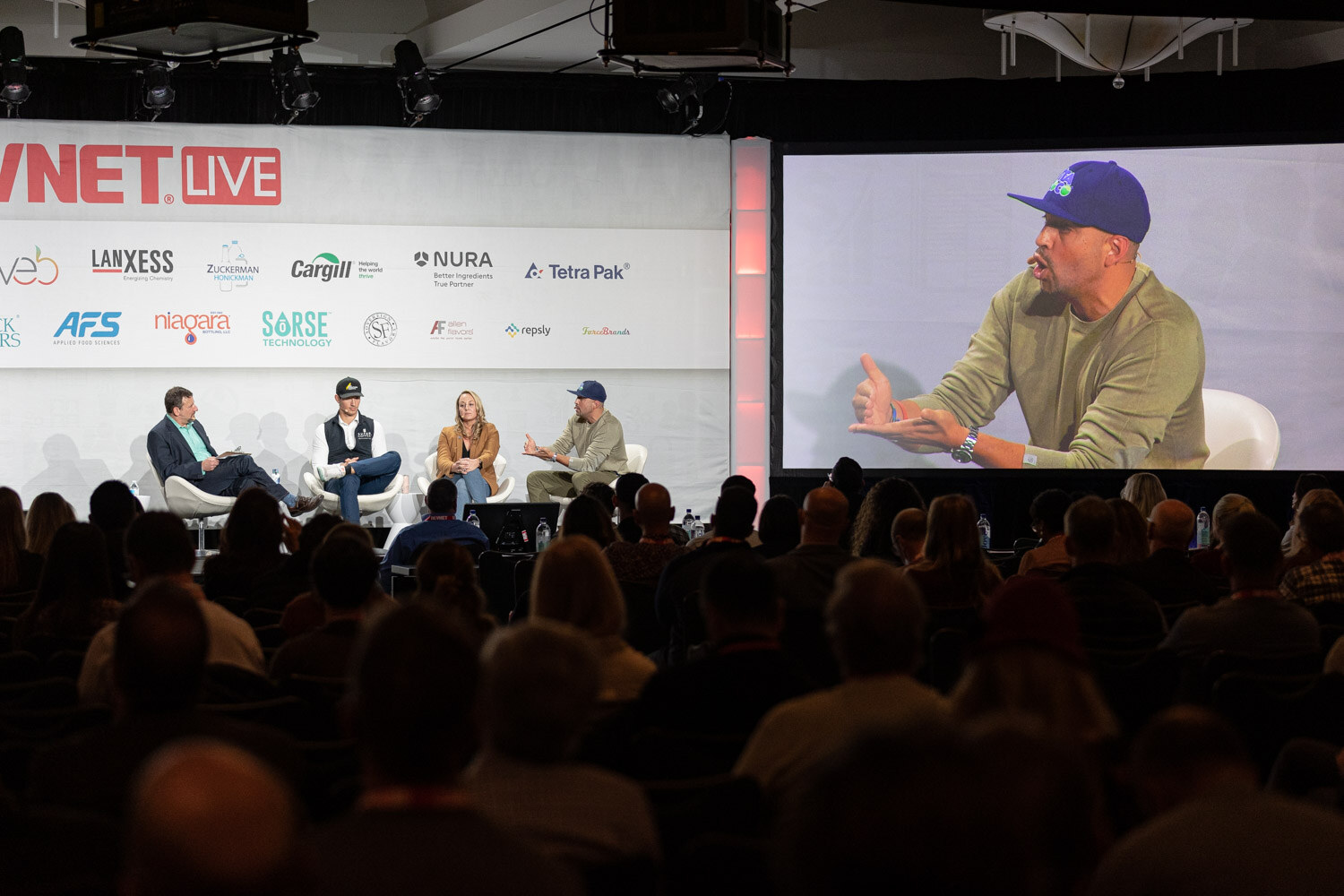 A wide view of four people on a stage under a BevNET Live banner, with a large screen at right showing a man in a blue cap, and the audience in the foreground.