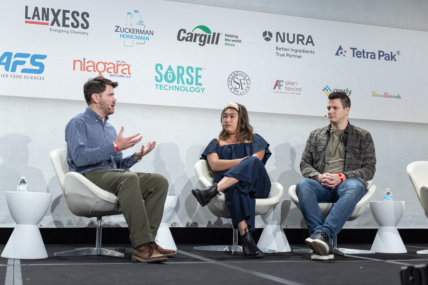 A man in a blue shirt gesturing, a woman in a navy jumpsuit and a man in a camouflage jacket sit in white chairs in front of a sponsor wall.