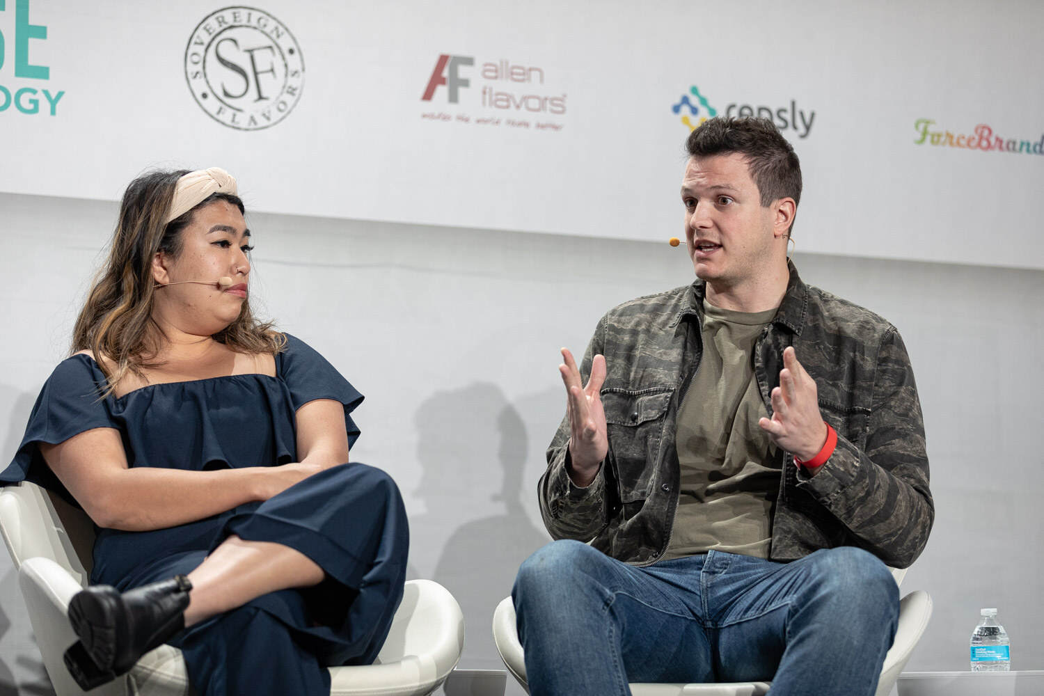 A woman in a navy jumpsuit and headband and a man in a camouflage jacket gesturing with both hands sit in white chairs.