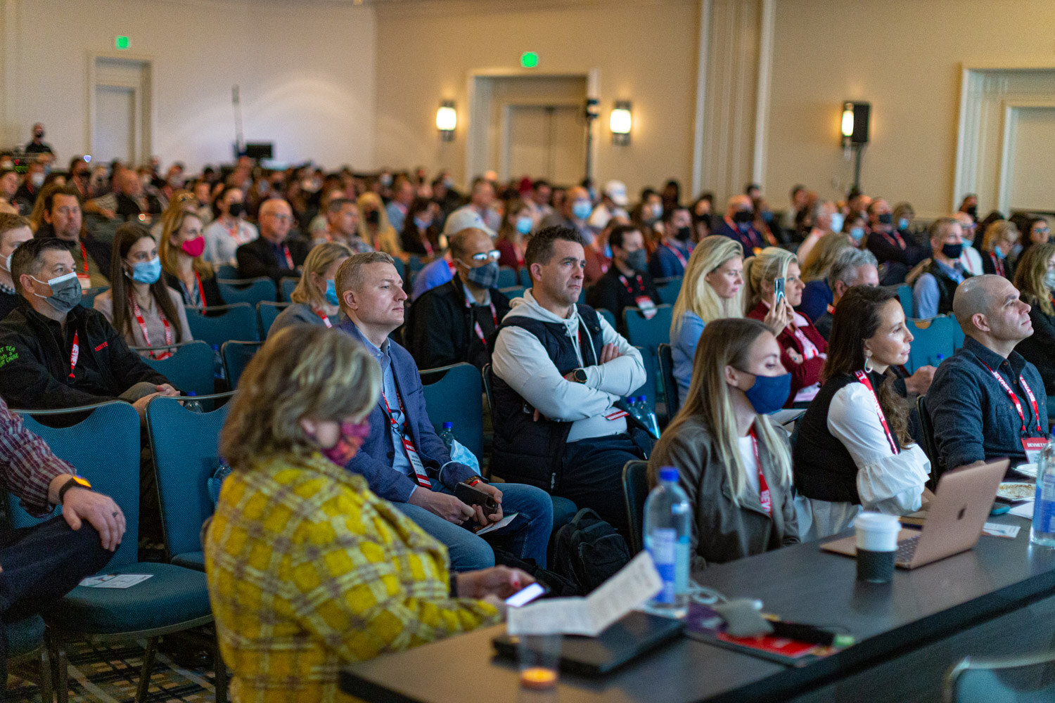 A crowded ballroom of seated attendees wearing lanyards, some in face masks, watching a stage out of view.