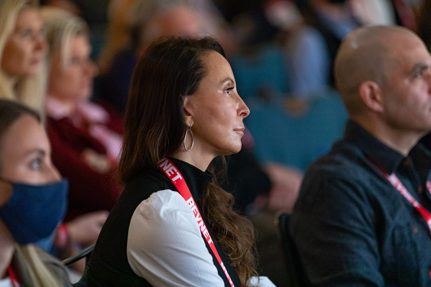 A woman with long dark hair and a BevNET lanyard in profile with a masked attendee and a bald man blurred in the background.