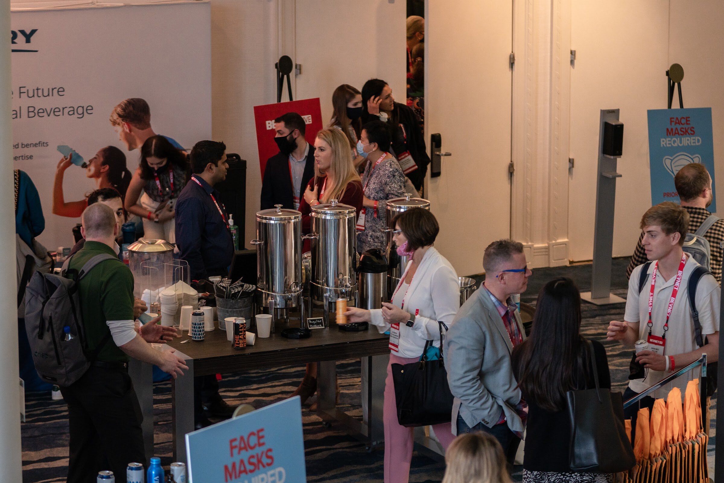 A high-angle view of attendees near coffee urns on a blue patterned carpet, with Face Masks Required signs and a Kerry banner in the background.