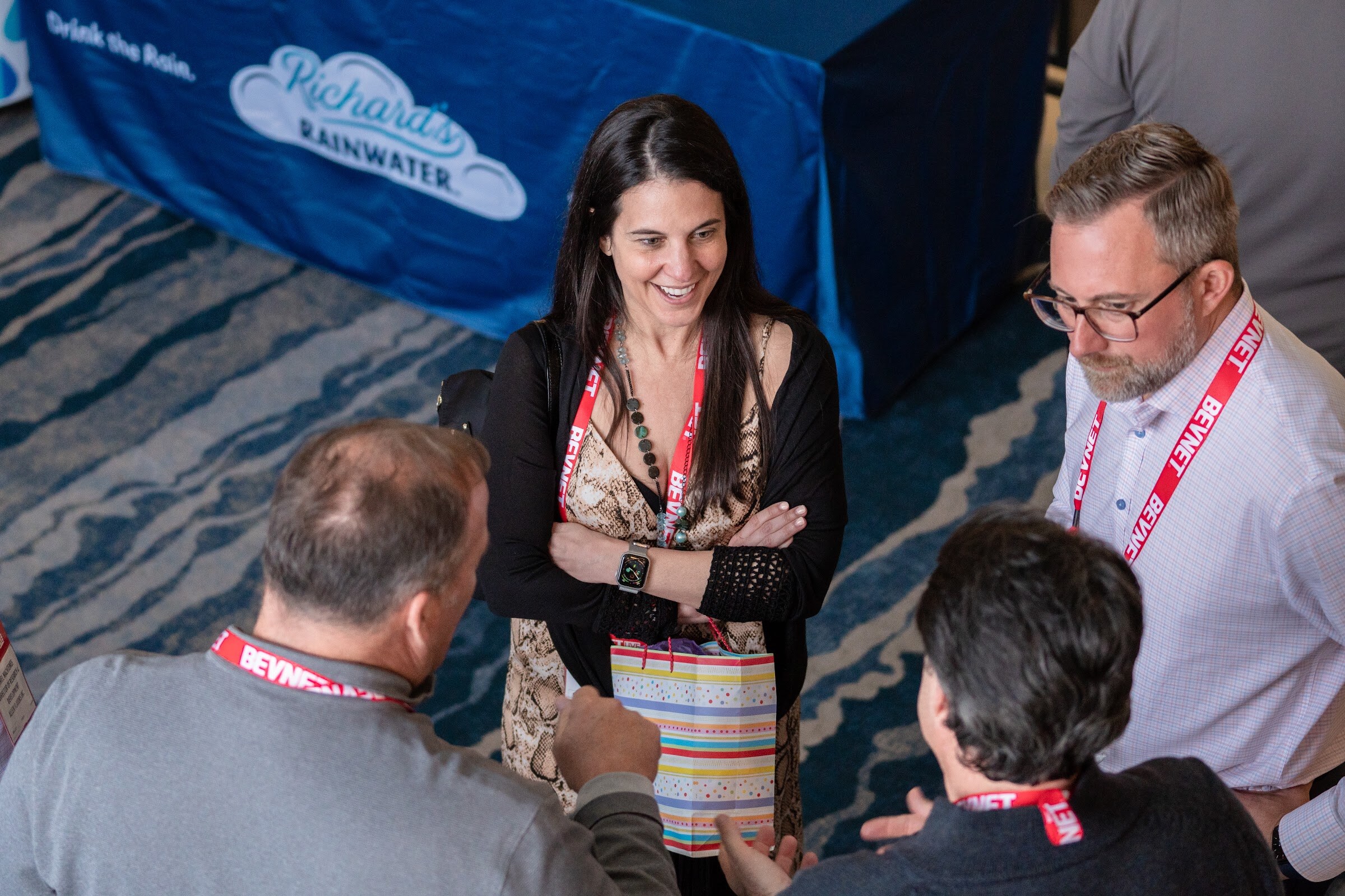 A high-angle view of a smiling woman with a BevNET lanyard holding a gift bag and talking with two men, with a blue Richard's Rainwater tablecloth behind them.