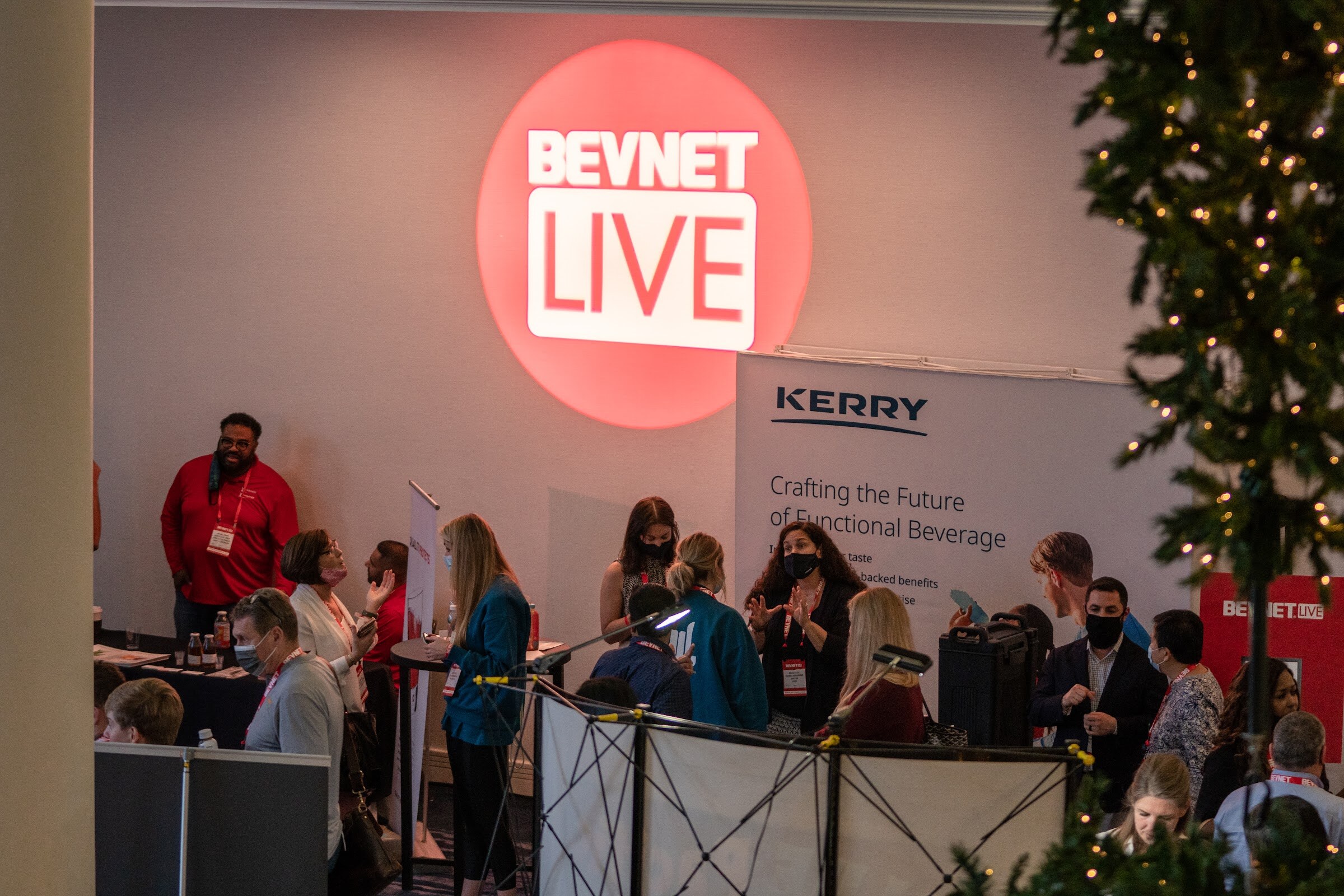 A view down on a busy expo area with a round red BevNET Live logo sign on the wall above a Kerry booth, with a Christmas tree at right.