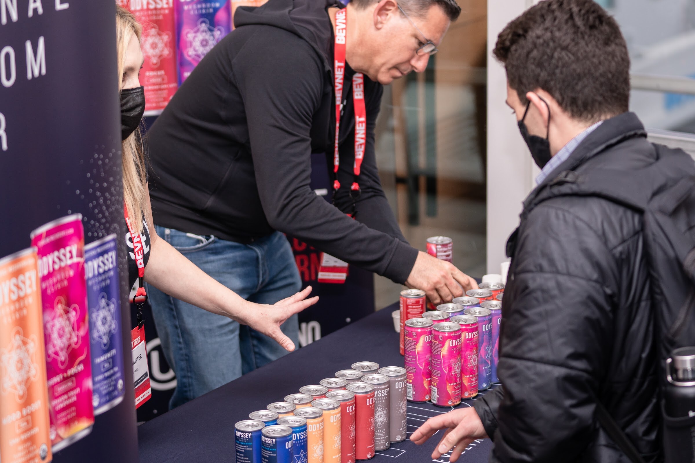 A man in a black hoodie leans over a navy table lined with rows of colorful Odyssey cans while another attendee in a puffer jacket looks on.
