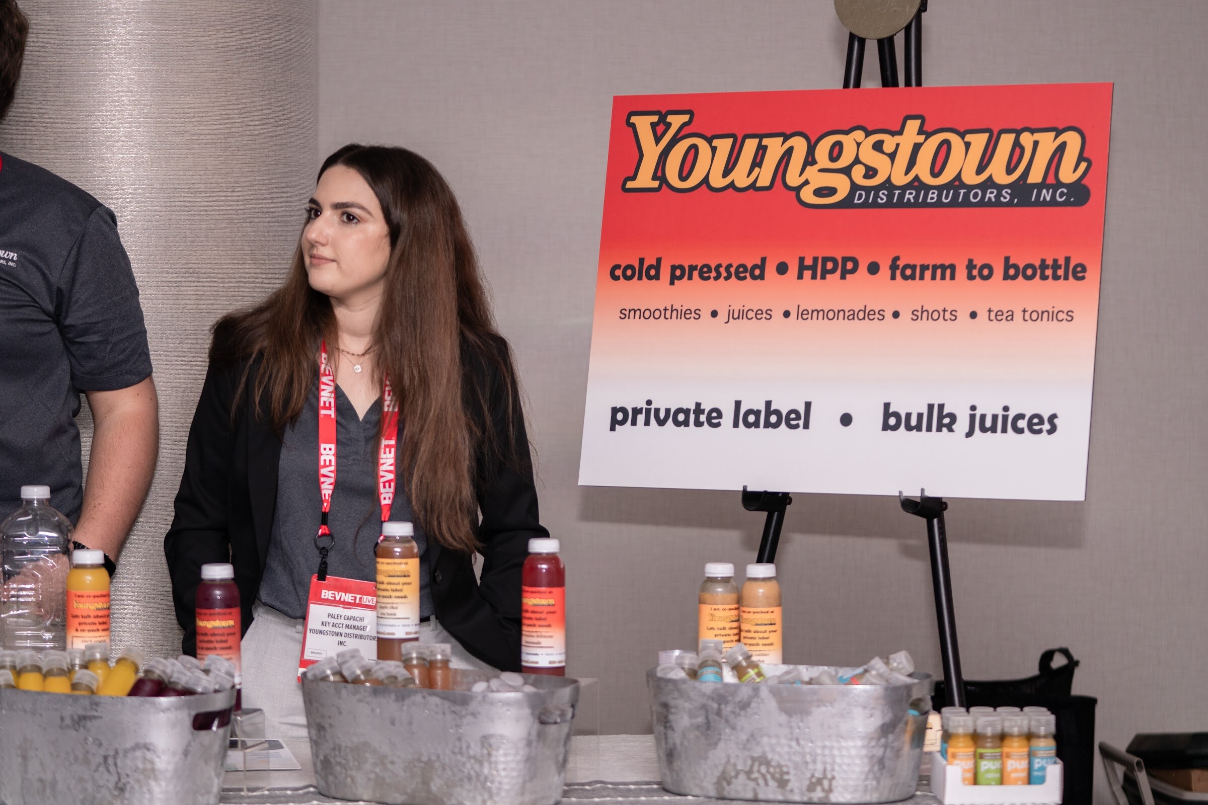 A woman with long brown hair and a BevNET lanyard stands behind a table of bottled juices in ice buckets next to a sign reading Youngstown Distributors, Inc. private label bulk juices.