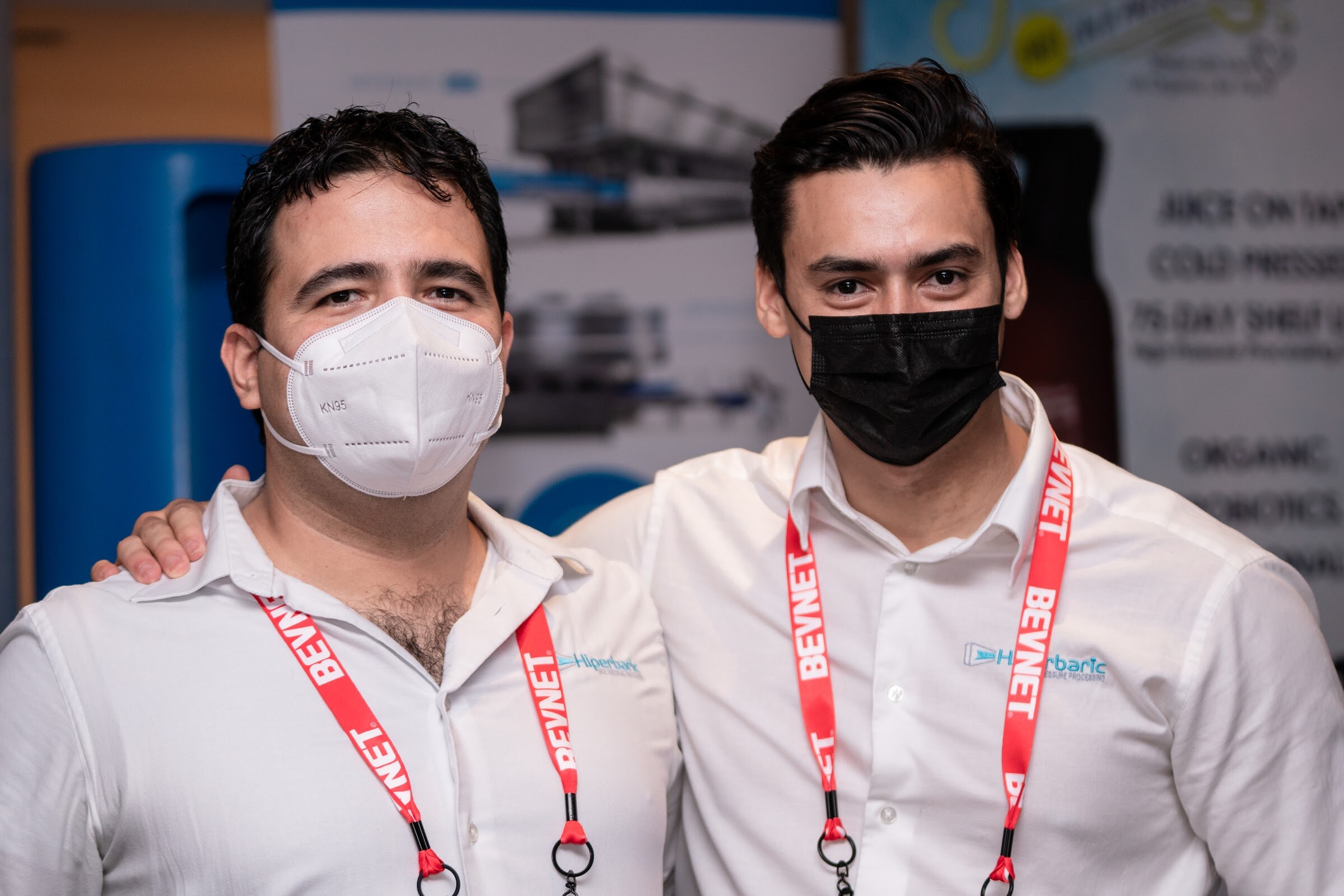 Two men in white collared shirts and face masks wearing red BevNET lanyards stand shoulder to shoulder in front of a booth display.