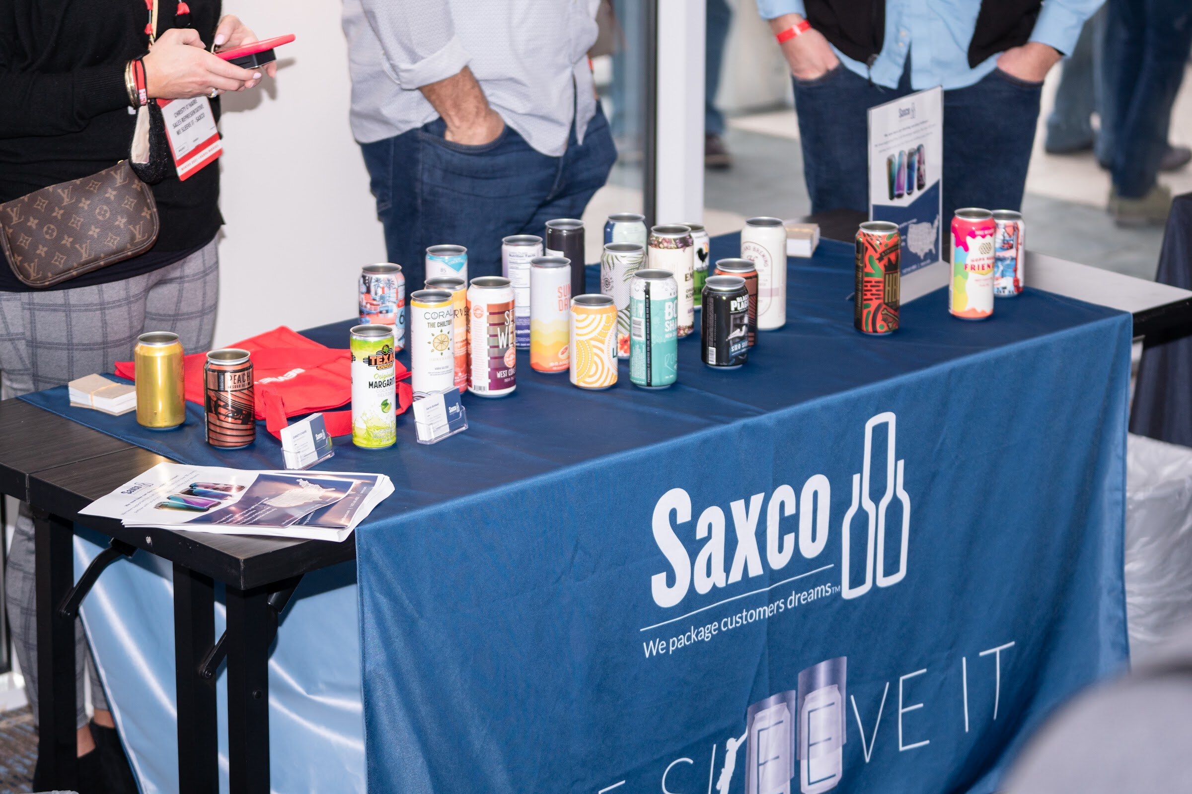 A navy tablecloth reading Saxco We package customers dreams covers a table with dozens of small beverage cans and printed brochures.