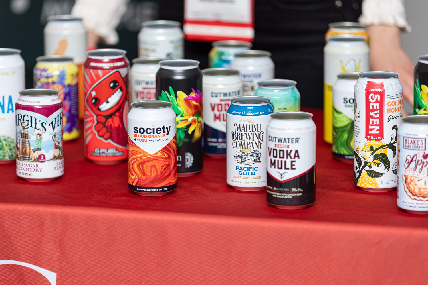 A tight view of many decorated cans, including Society, Malibu Brewing Company, Cutwater Vodka Mule and Seven Tea, on a red tablecloth.
