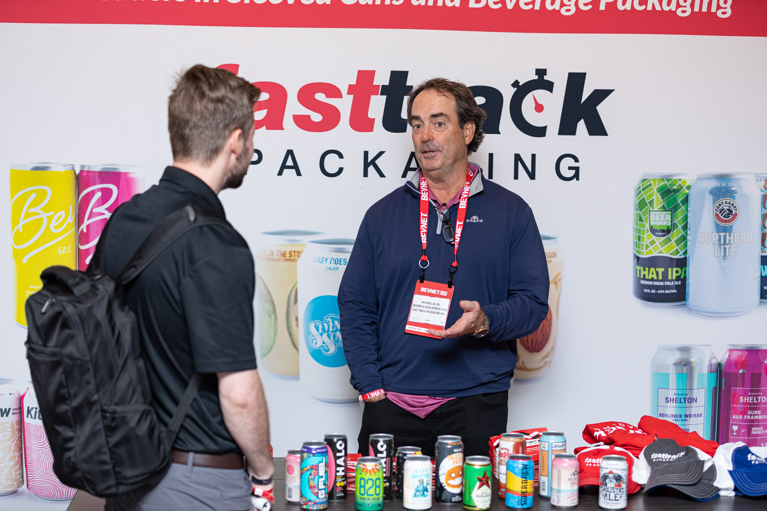 A man in a navy quarter-zip with a BevNET lanyard speaks to an attendee carrying a backpack in front of a Fasttrack Packaging banner, with cans along the table.