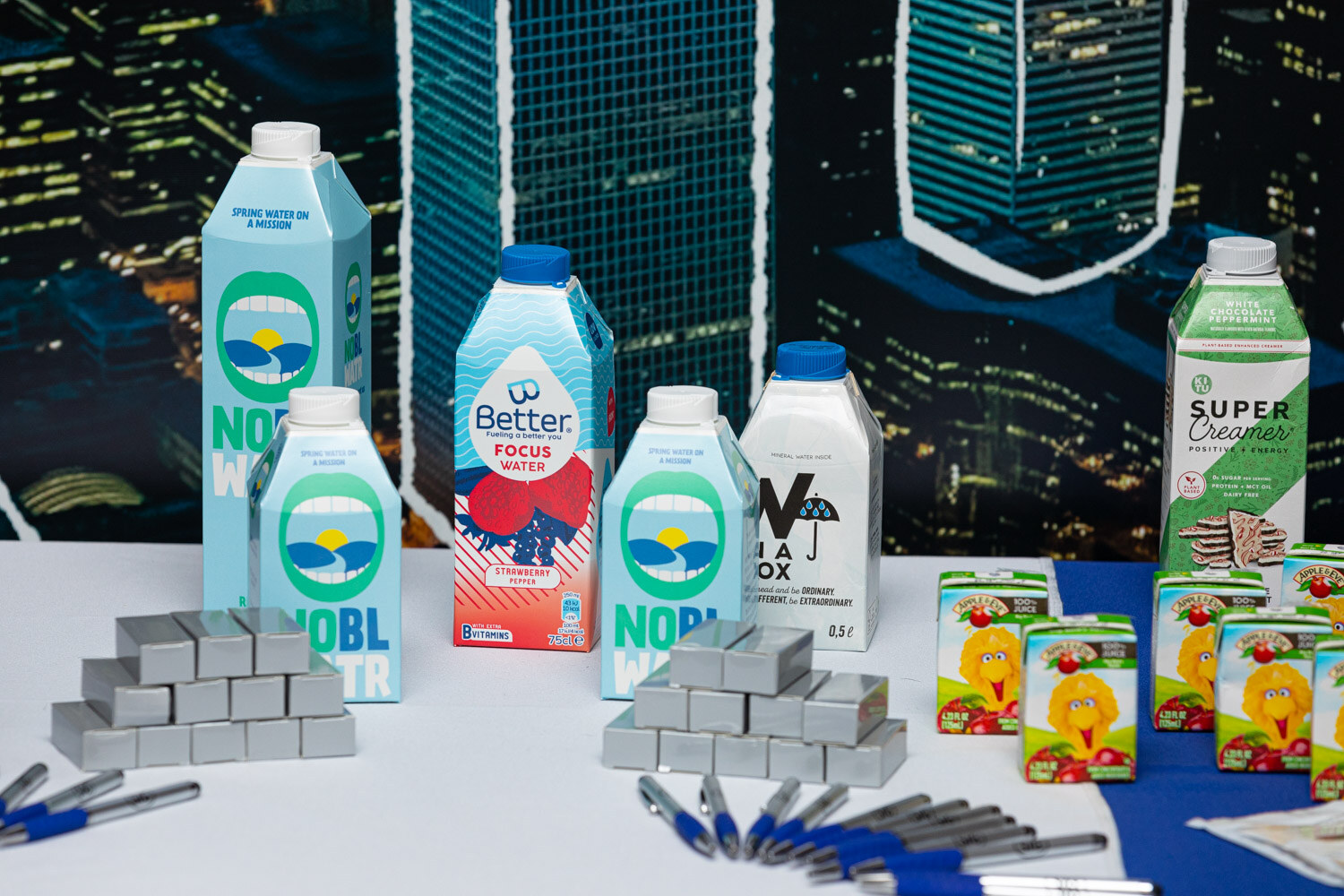 Tall cartons including Nobl, Better and Super Creamer stand on a white table beside small juice boxes and pens in front of a city skyline backdrop.