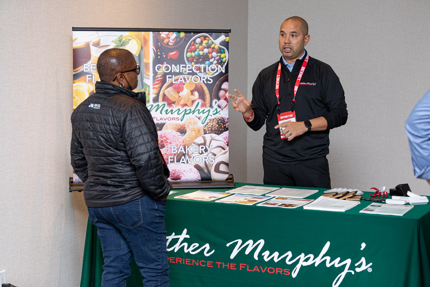 A man in a black quarter-zip with a BevNET lanyard speaks to an attendee in a puffer jacket in front of a Mother Murphy's table with a green tablecloth and confection flavors banner.