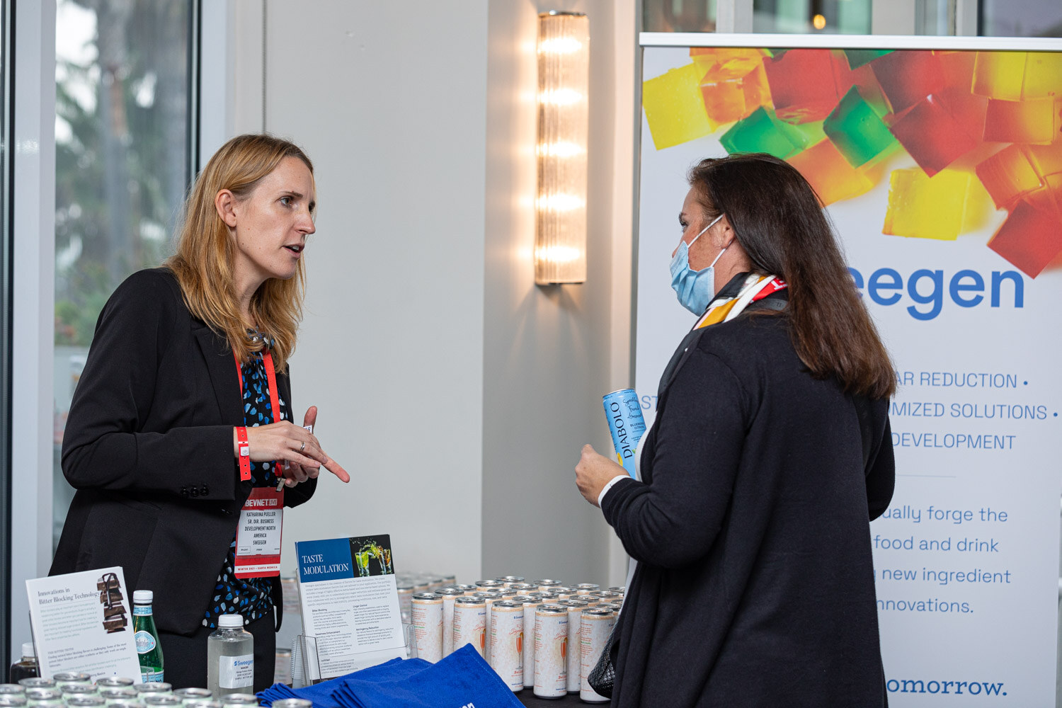 A blonde woman in a black blazer talks with a woman in a face mask in front of a banner reading Sweegen, with cans and brochures on the table.