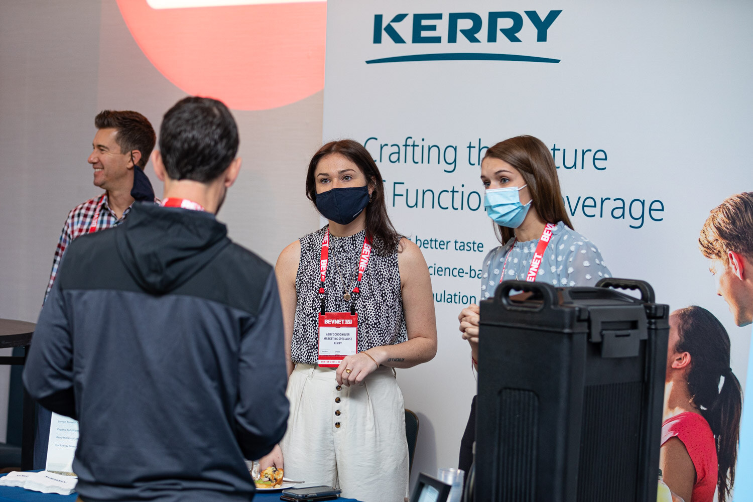 Several people wearing BevNET lanyards stand in front of a banner reading Kerry crafting the future of functional beverage, with a black beverage dispenser in the foreground.