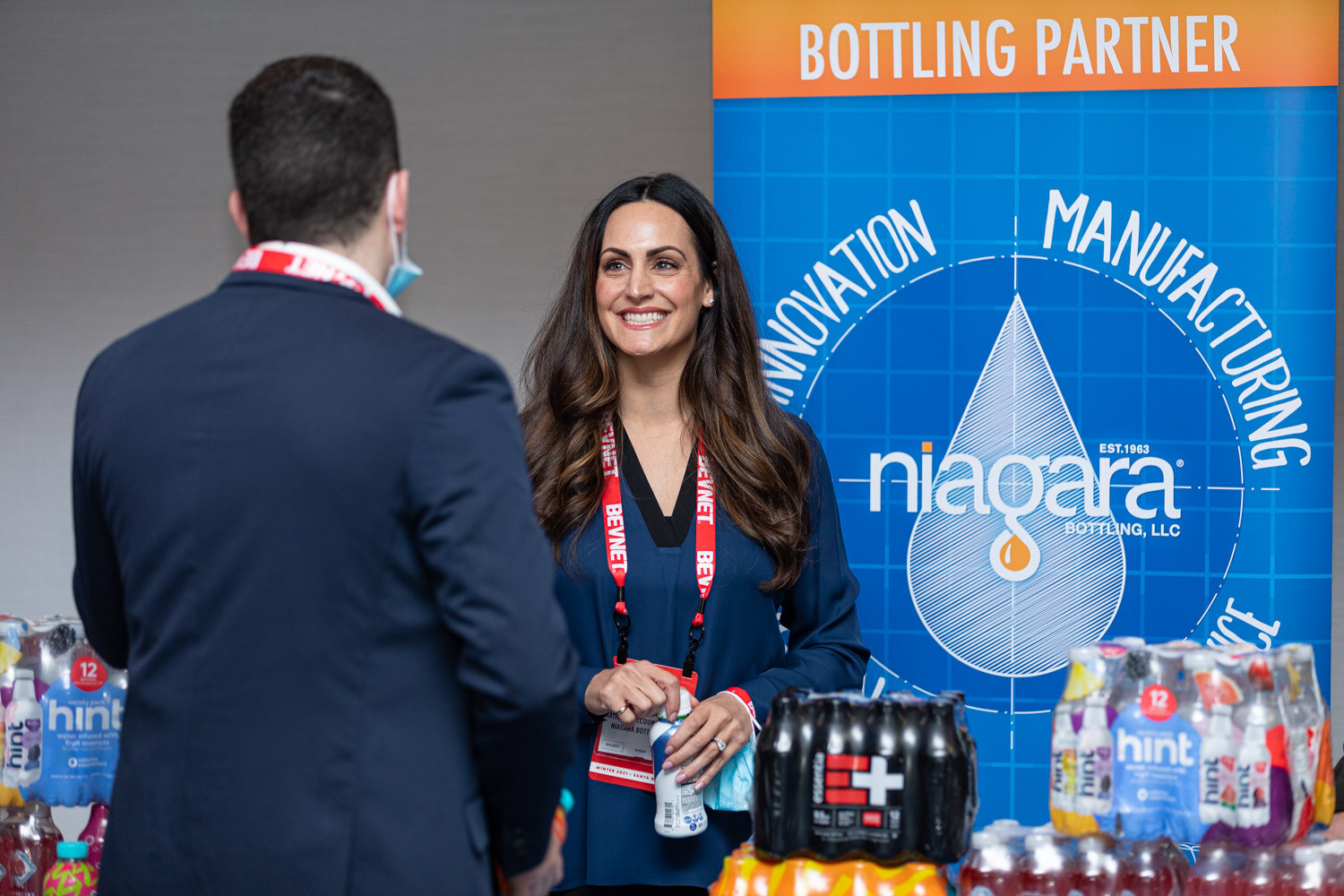 A woman with long dark hair and a BevNET lanyard stands in front of a blue Niagara Bottling banner with stacks of hint and E+ beverages on the table.