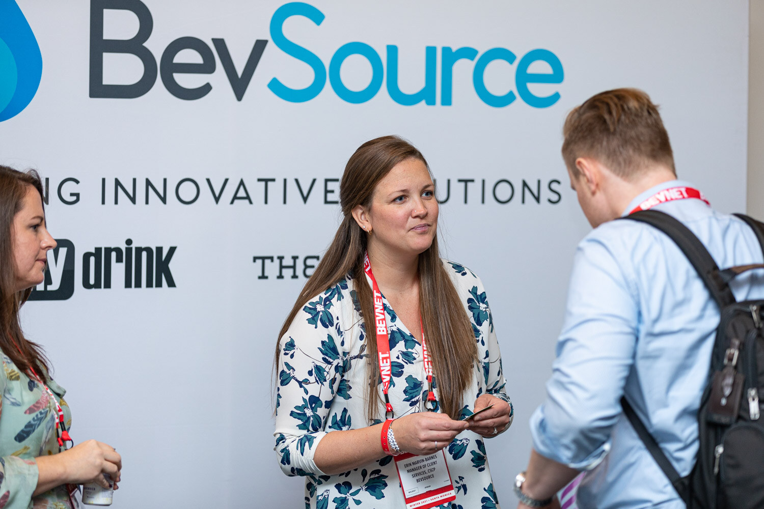 A woman in a floral blouse with a BevNET lanyard speaks with an attendee wearing a backpack in front of a large BevSource sign.