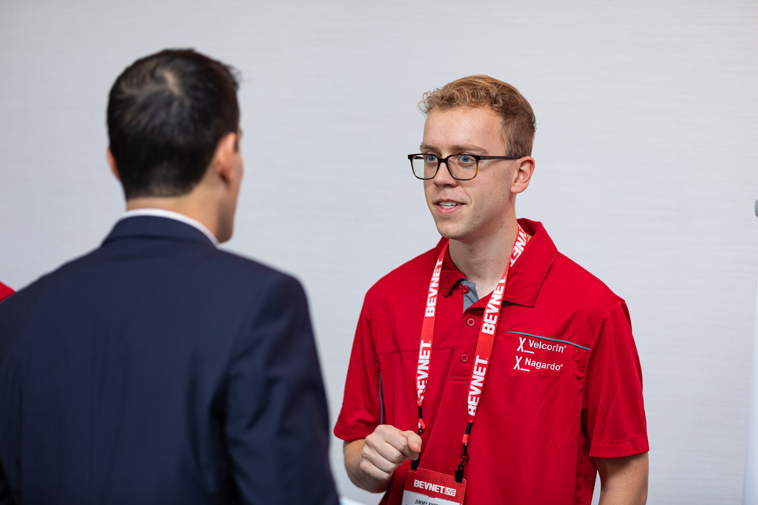 A young man with glasses in a red Velcorin Nagardo polo and BevNET lanyard speaks with a man in a dark suit in front of a white wall.