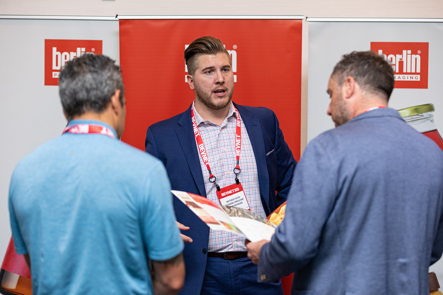 A man in a navy blazer holding a brochure speaks to two attendees in front of red Berlin Packaging banners.