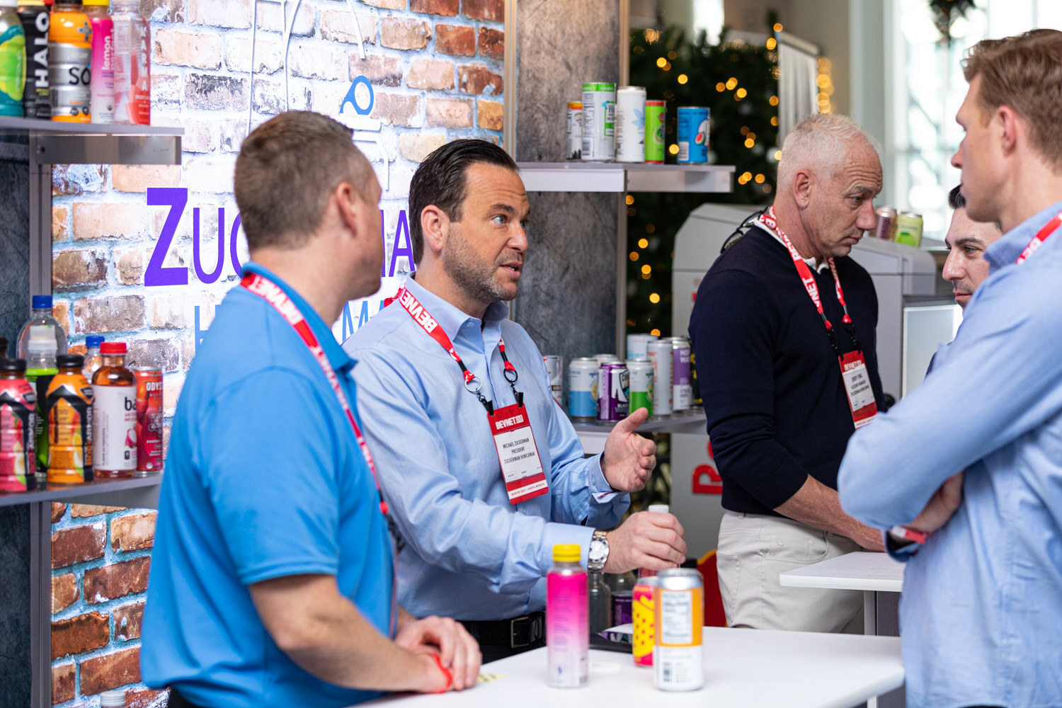 Five men with BevNET lanyards talk around a white table in front of a brick wall with Zuckerman lettering and a shelf of drinks.