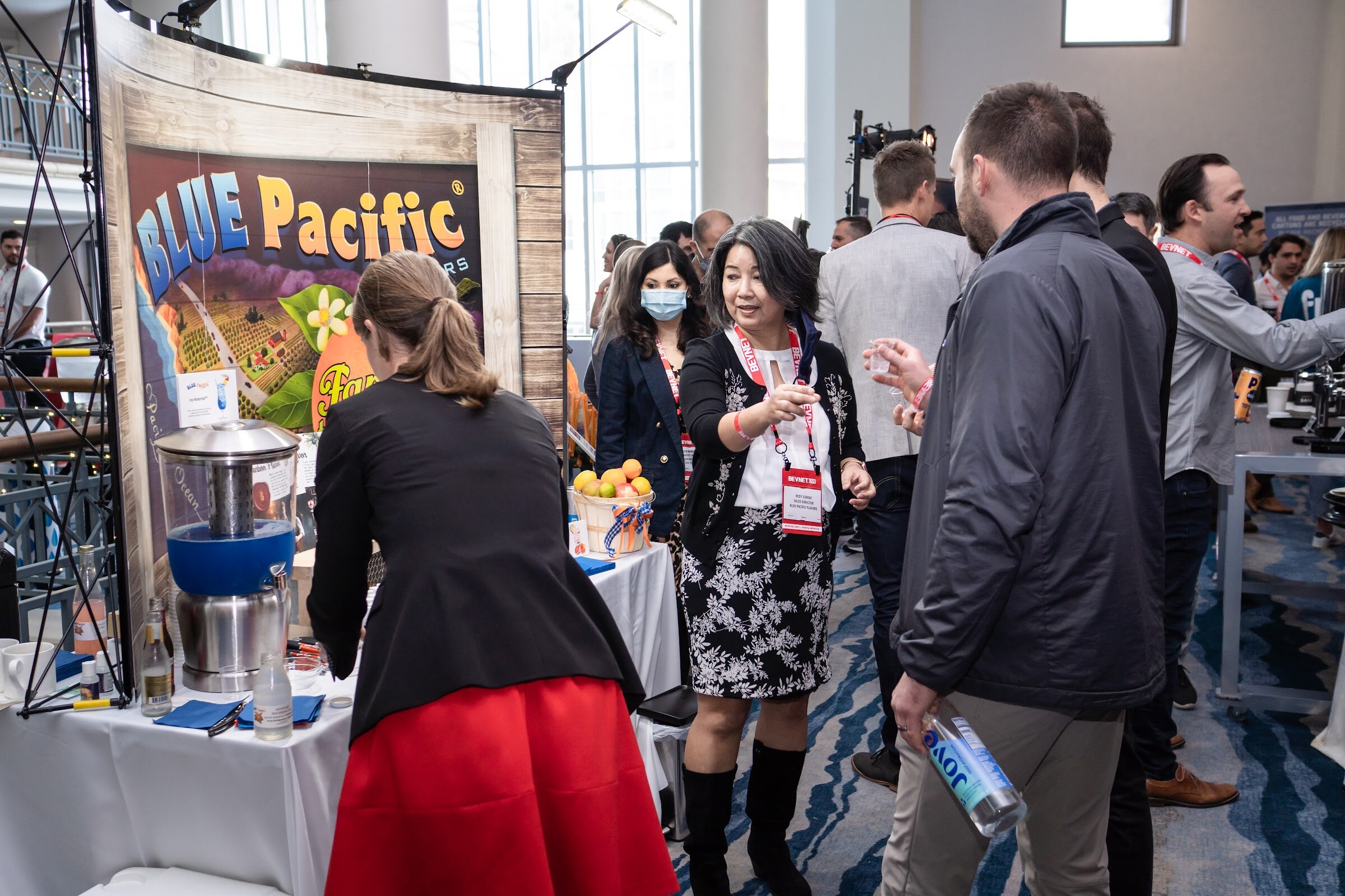 A crowded floor with attendees wearing lanyards around a Blue Pacific Flavors backdrop, a juicer on the table and a woman in a red skirt in the foreground.