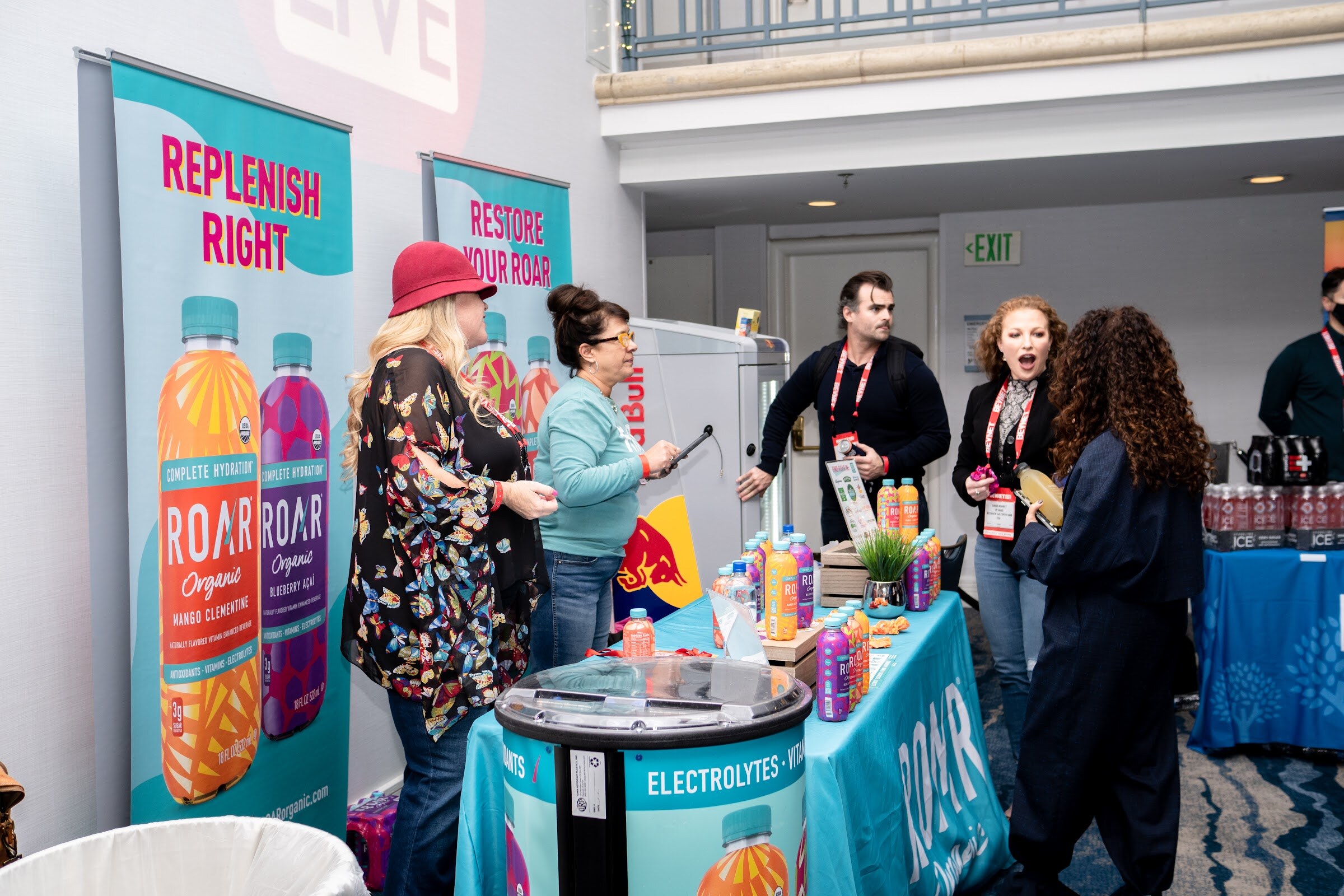 Several people in front of teal Roar Organic banners reading Replenish Right and Restore Your Roar, around a table of bottles and a hat-wearing exhibitor.