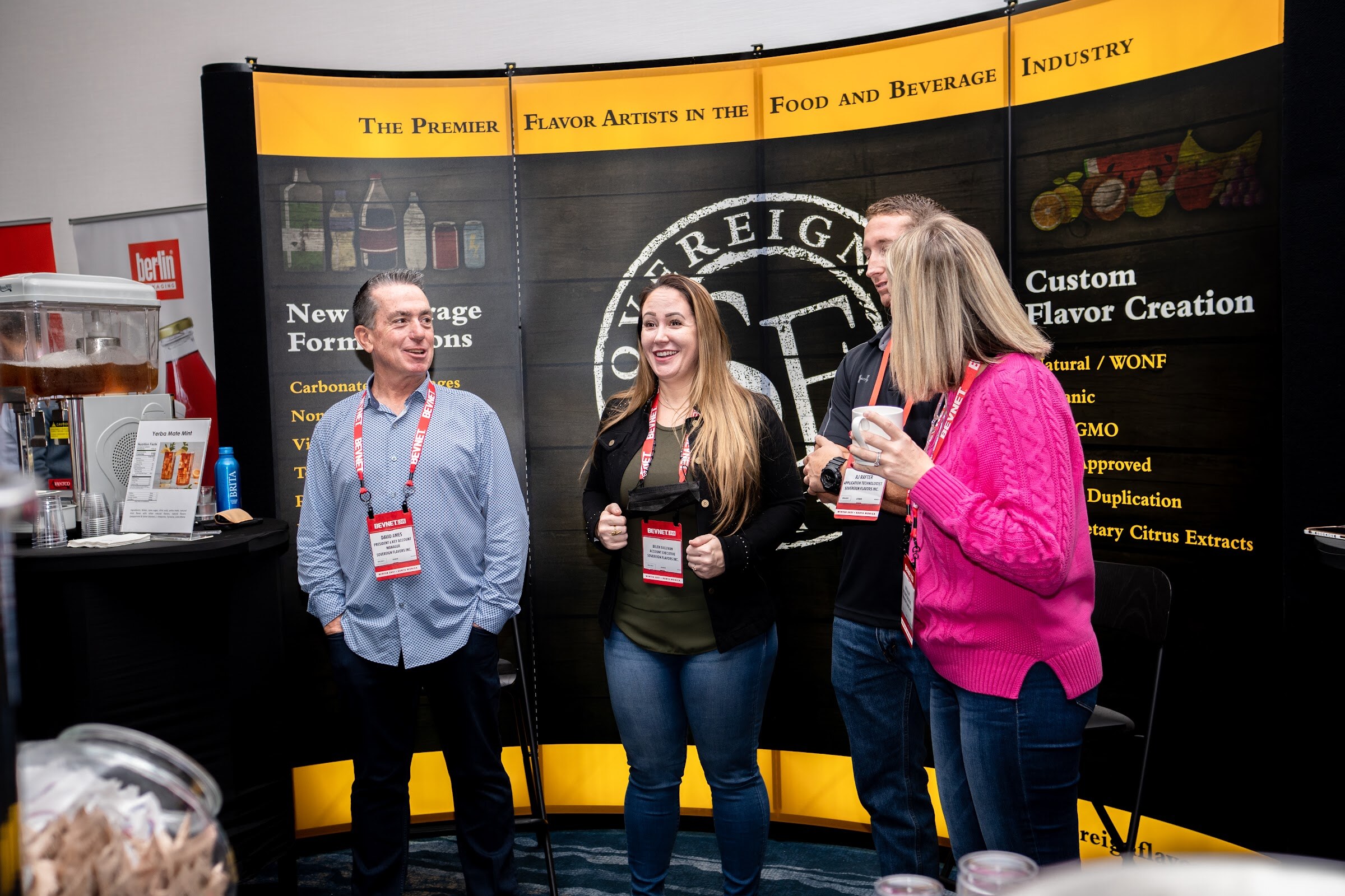 Four people wearing BevNET lanyards stand smiling in front of a black and yellow curved display reading The Premier Flavor Artists in the Food and Beverage Industry.