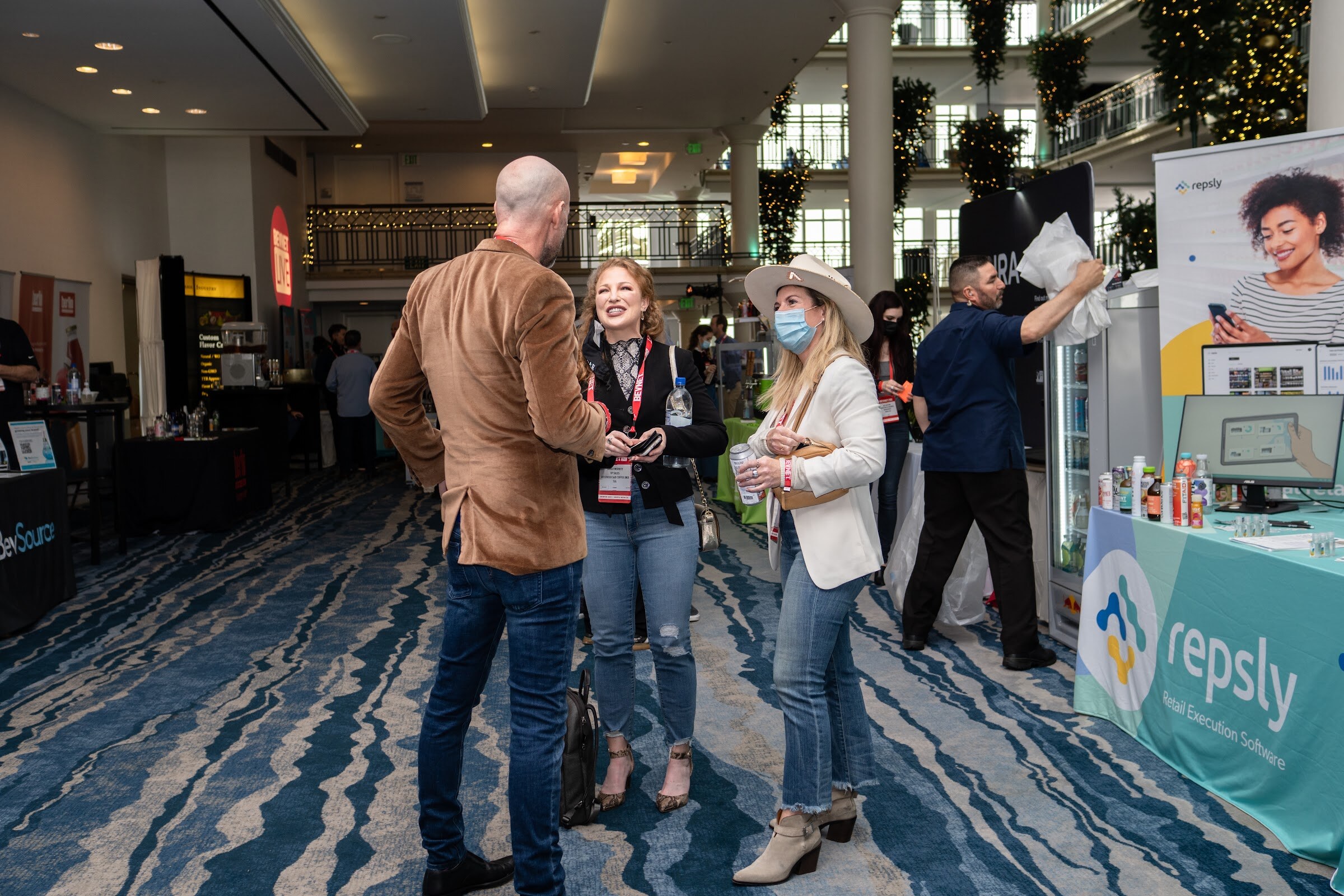 Three attendees, including a woman in a white blazer and cowboy hat, stand in conversation on a blue wave-pattern carpet near a Repsly booth.