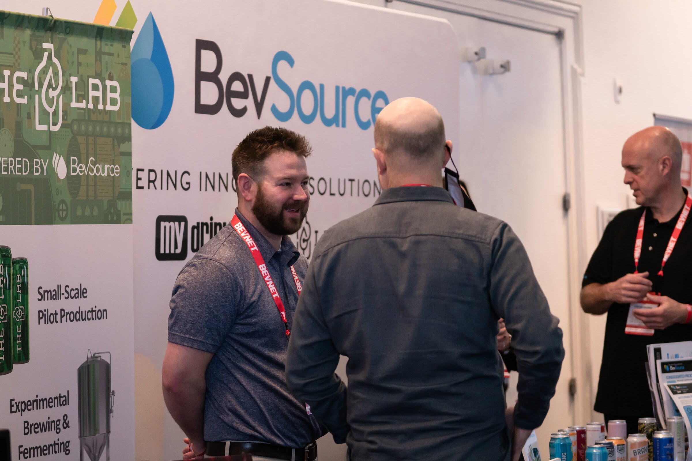 A bearded man in a gray polo and BevNET lanyard smiles at a bald man in a gray shirt in front of a BevSource and The Lab small-scale pilot production banner.