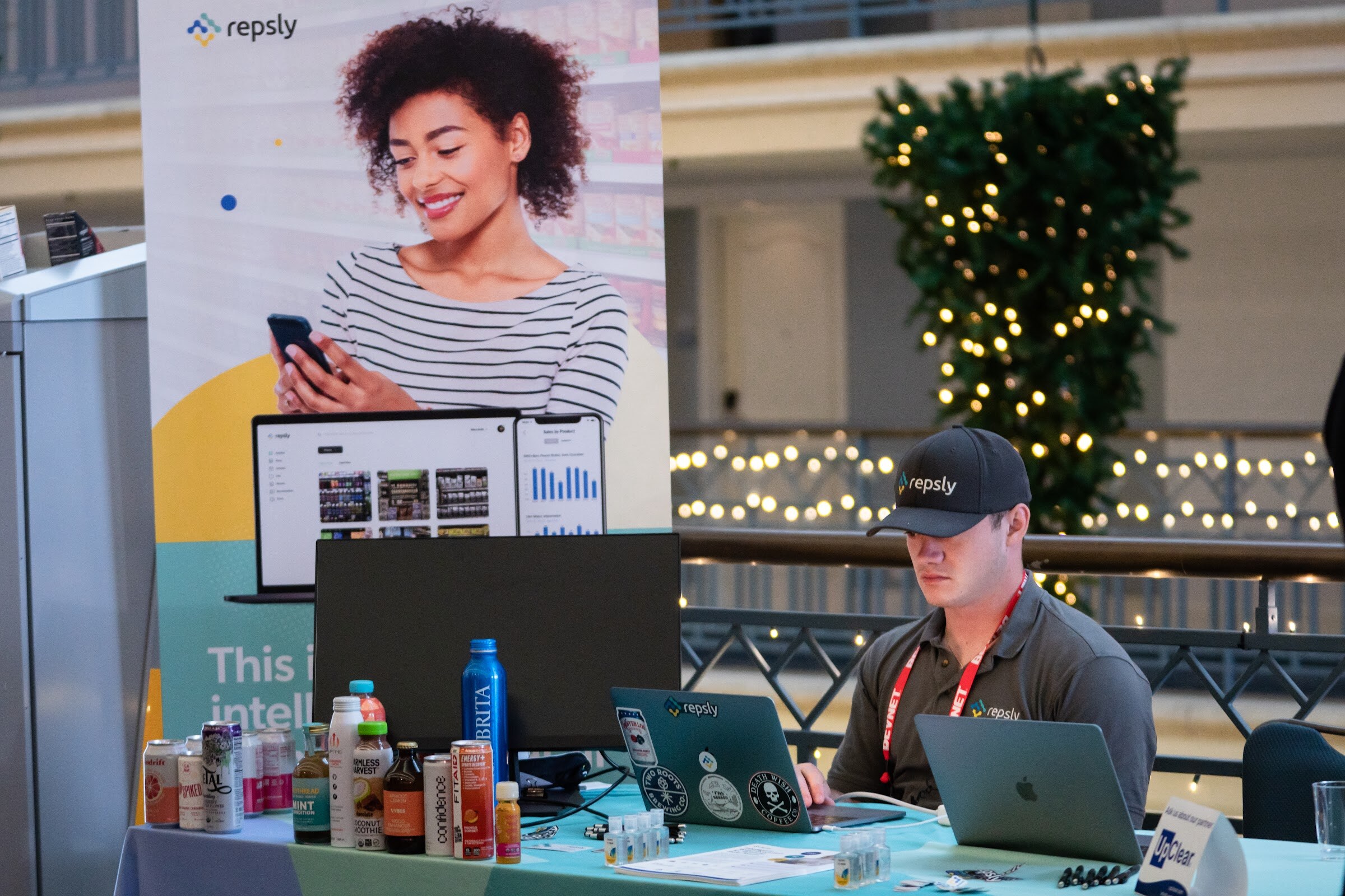A man in a Repsly cap and BevNET lanyard looks down at a laptop on a table with beverage cans, in front of a Repsly banner and a decorated Christmas tree.