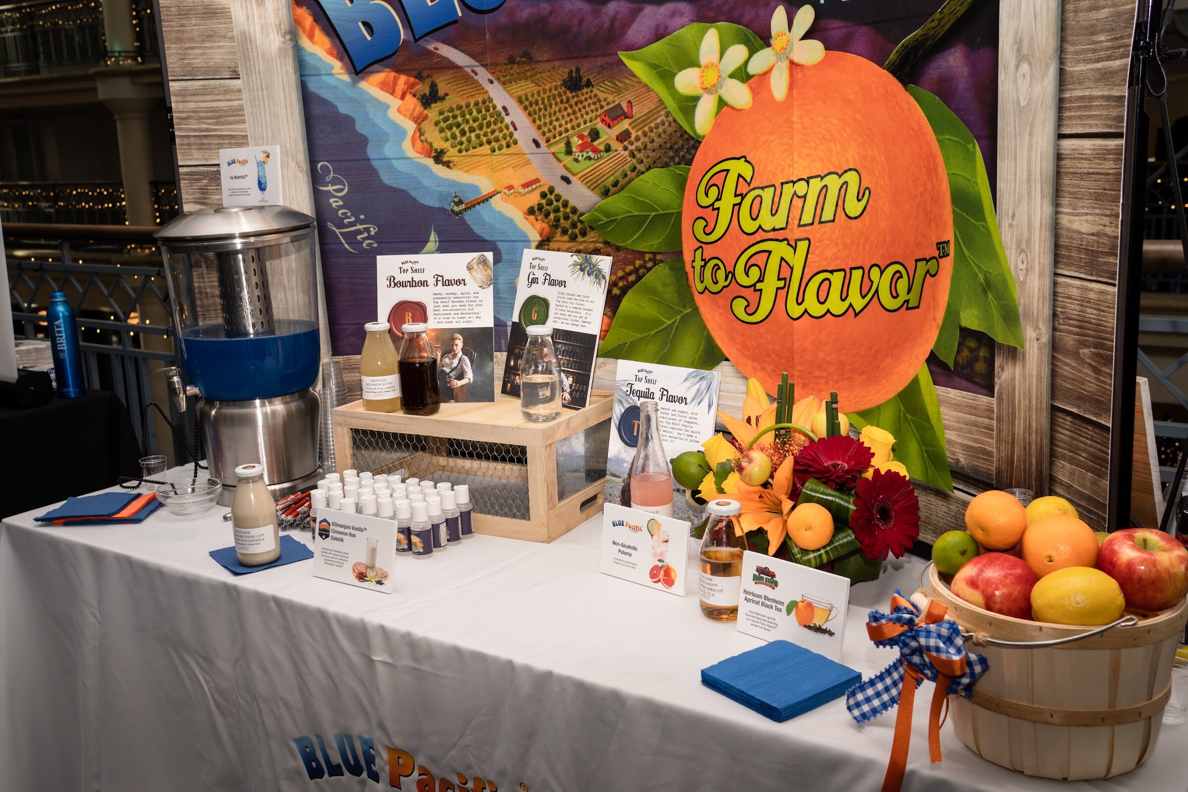 A white-clothed table with a juice dispenser, small bottles, a basket of citrus and a backdrop painted with the Farm to Flavor logo.