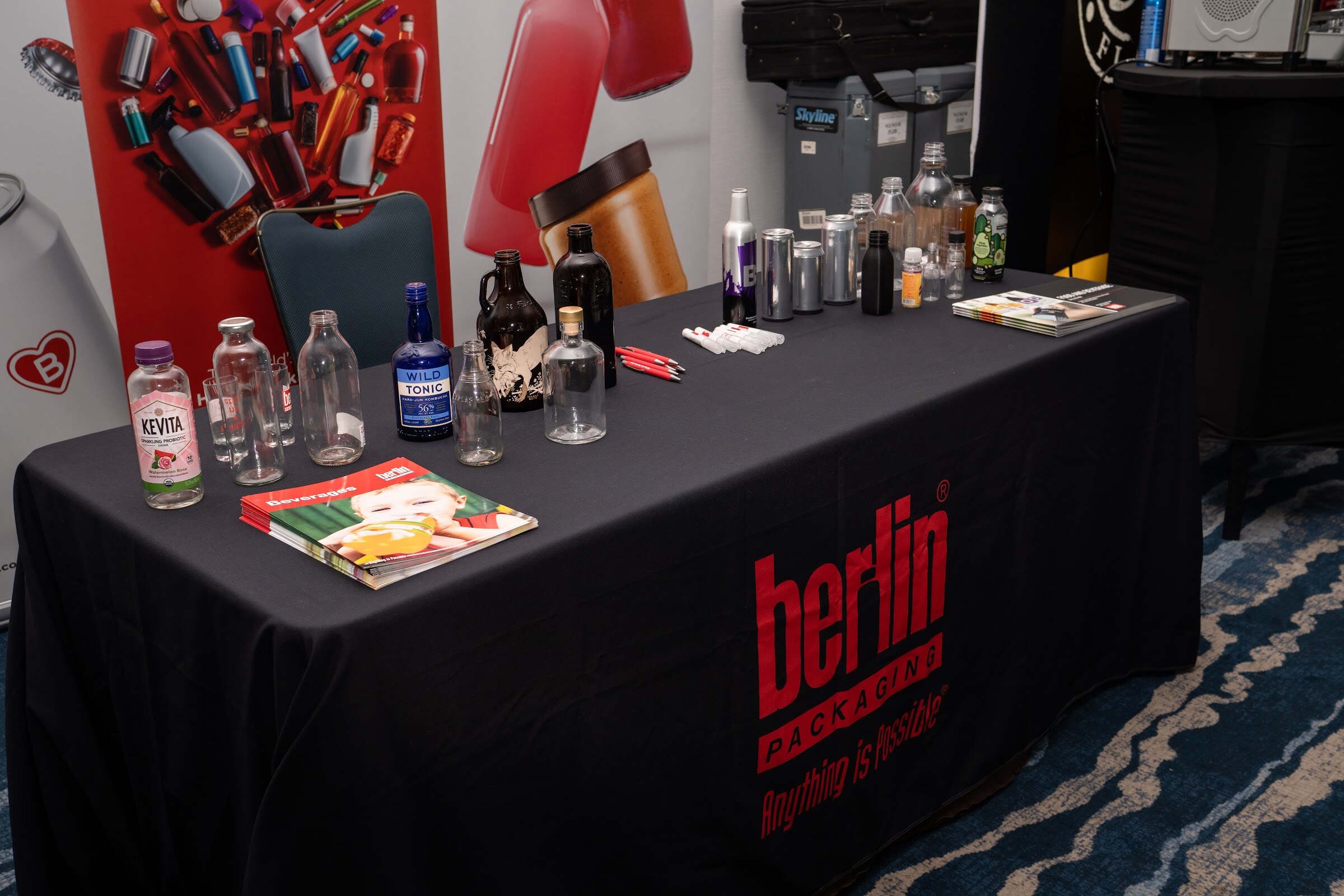 A black tablecloth printed with Berlin Packaging holds clear and amber bottles, a Kevita bottle and a brochure.