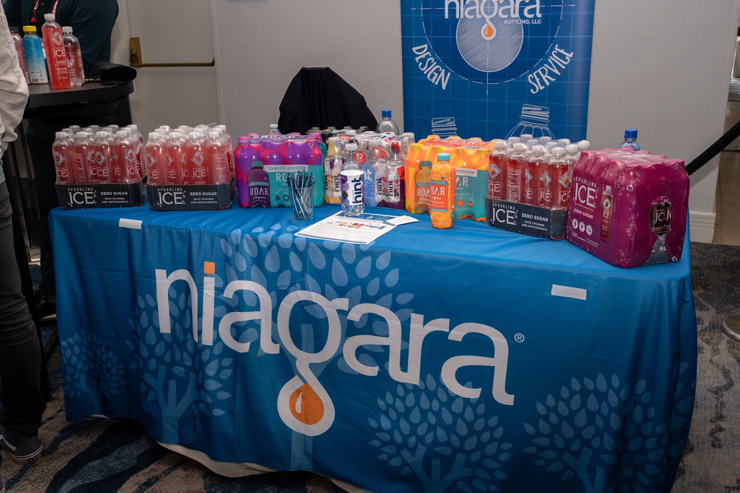 A blue tablecloth printed with the Niagara logo holds stacked cases of colorful sparkling and flavored waters in front of a blue banner.